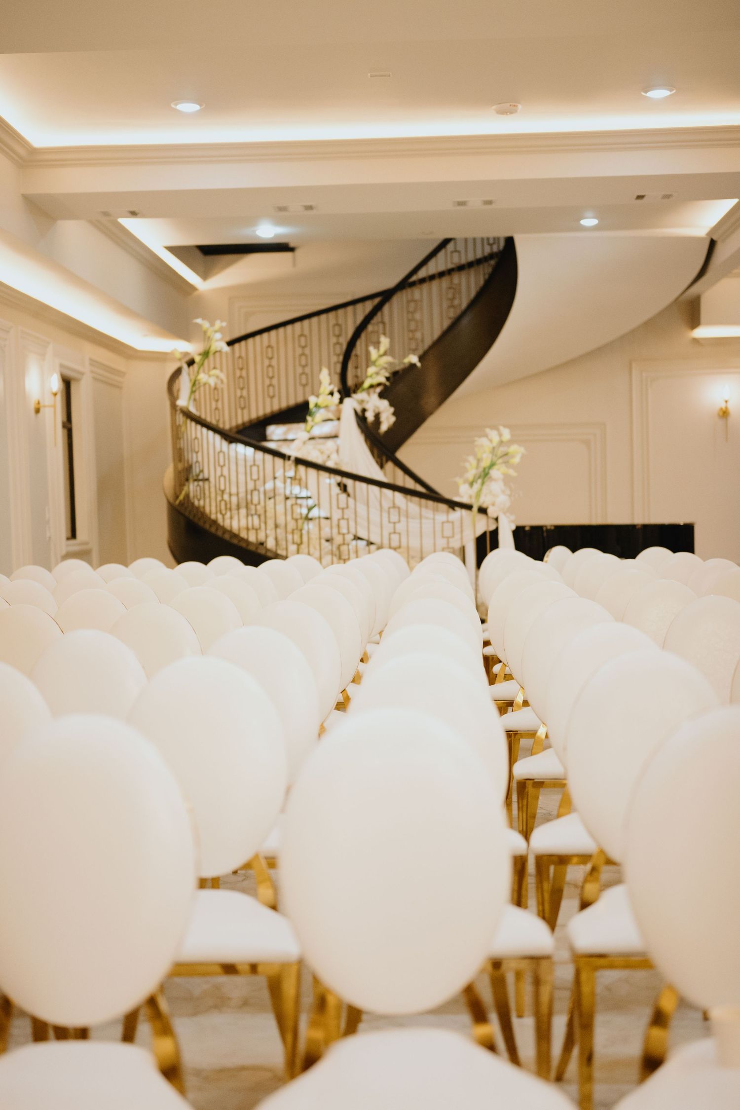 Rows of white oval-back chairs with gold legs face an elegant curved staircase decorated with white floral arrangements at a wedding venue.