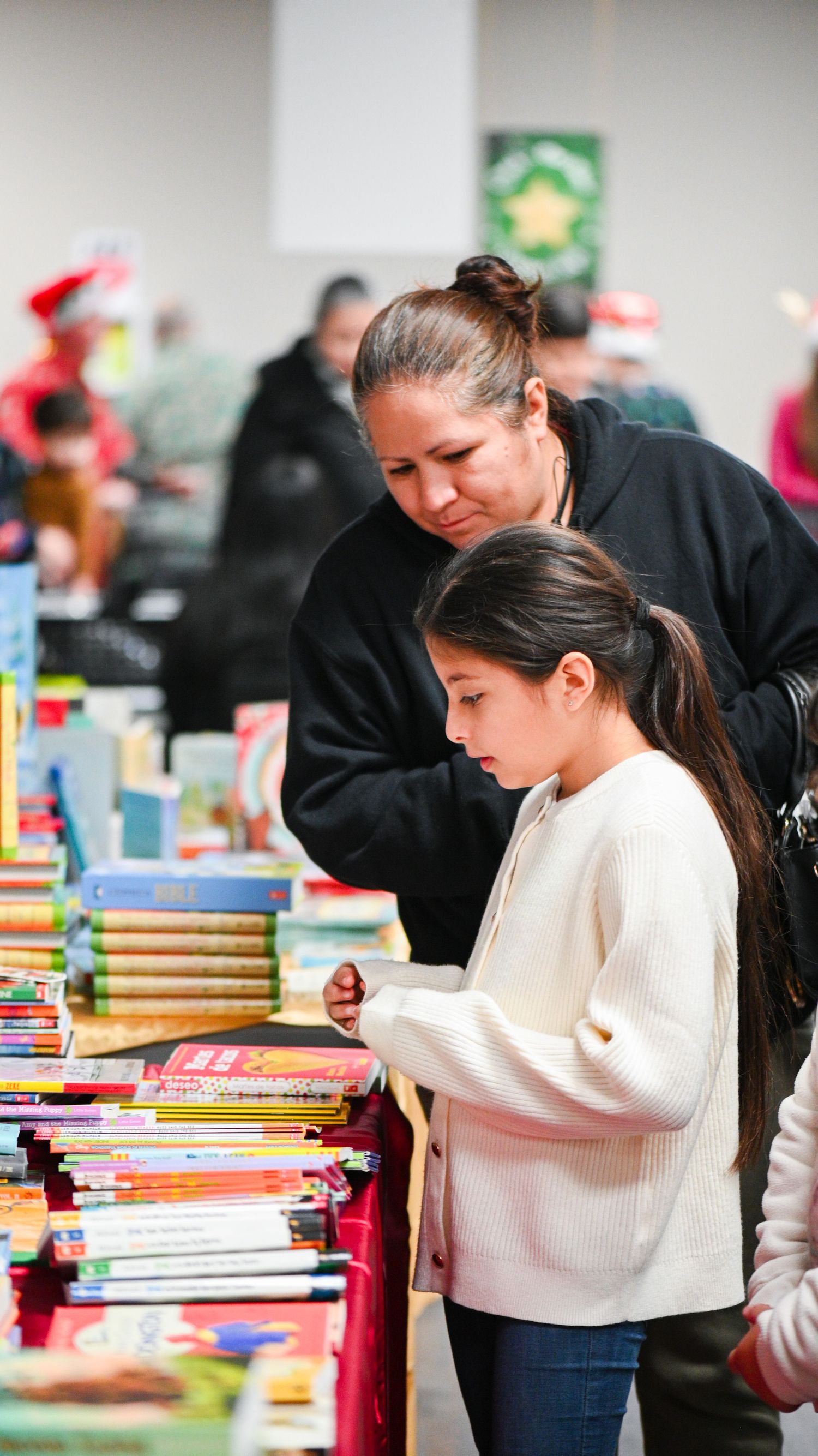 A woman and a young girl browse tables of children's books at a holiday book giveaway event.