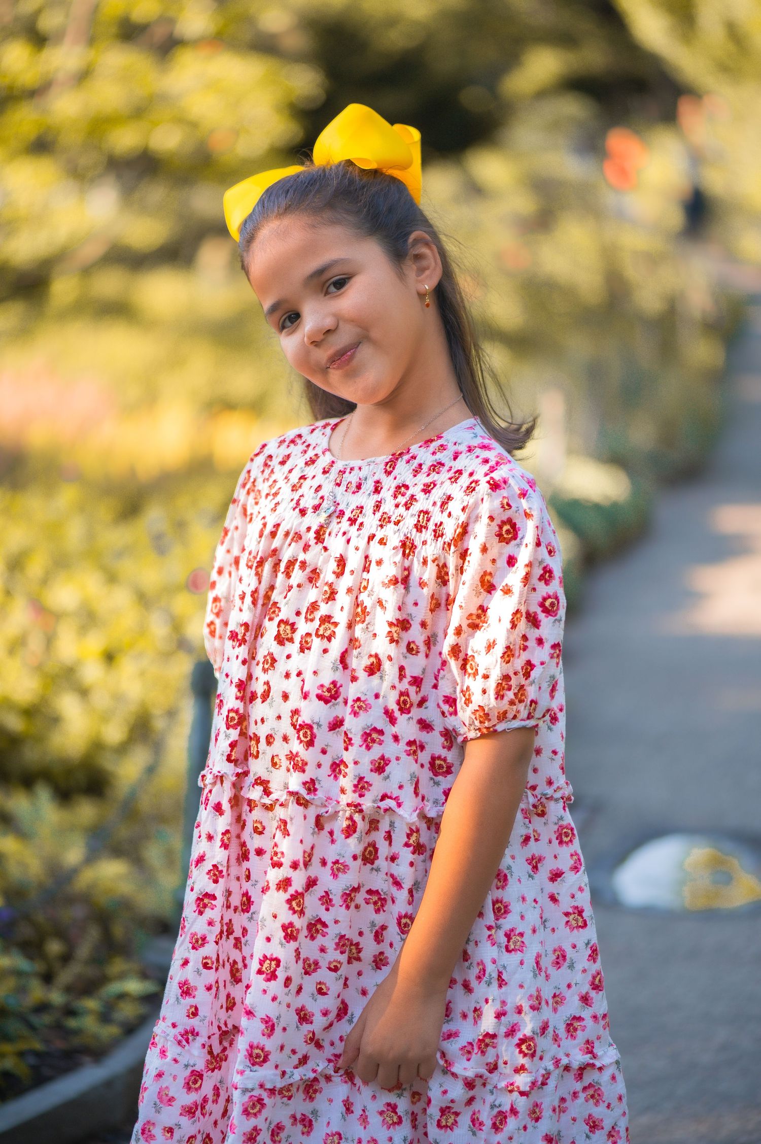 A young girl in a pink floral dress and yellow hair bow smiles for a portrait on a garden path in warm evening light.