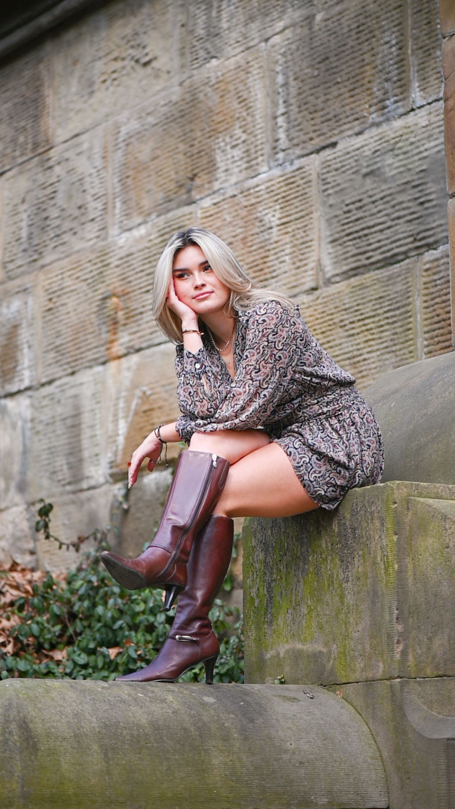 Blonde woman in a floral mini dress and tall brown leather boots sitting on a mossy stone ledge in front of a sandstone wall.