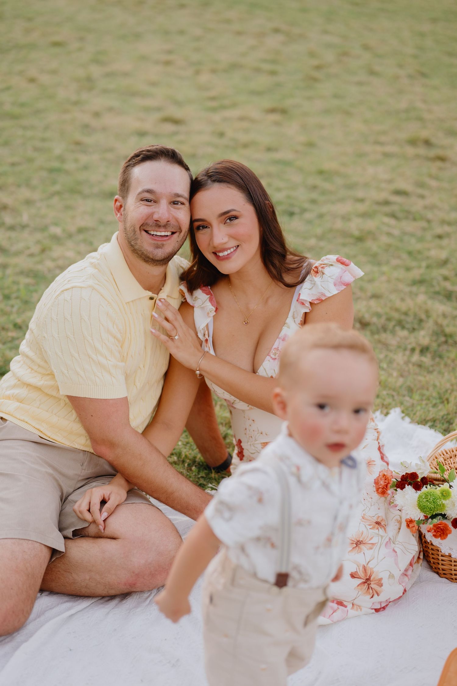 A smiling couple sits close together on a picnic blanket in a grassy park while their toddler son stands in front of them.