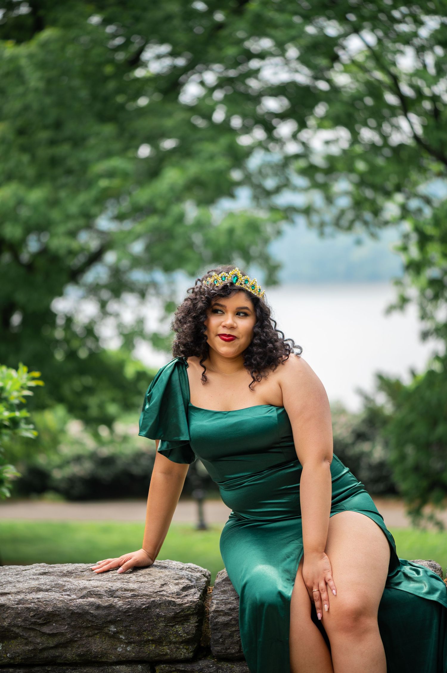 Woman with curly hair wears a green satin gown and jeweled crown while posing on a stone wall by the water.