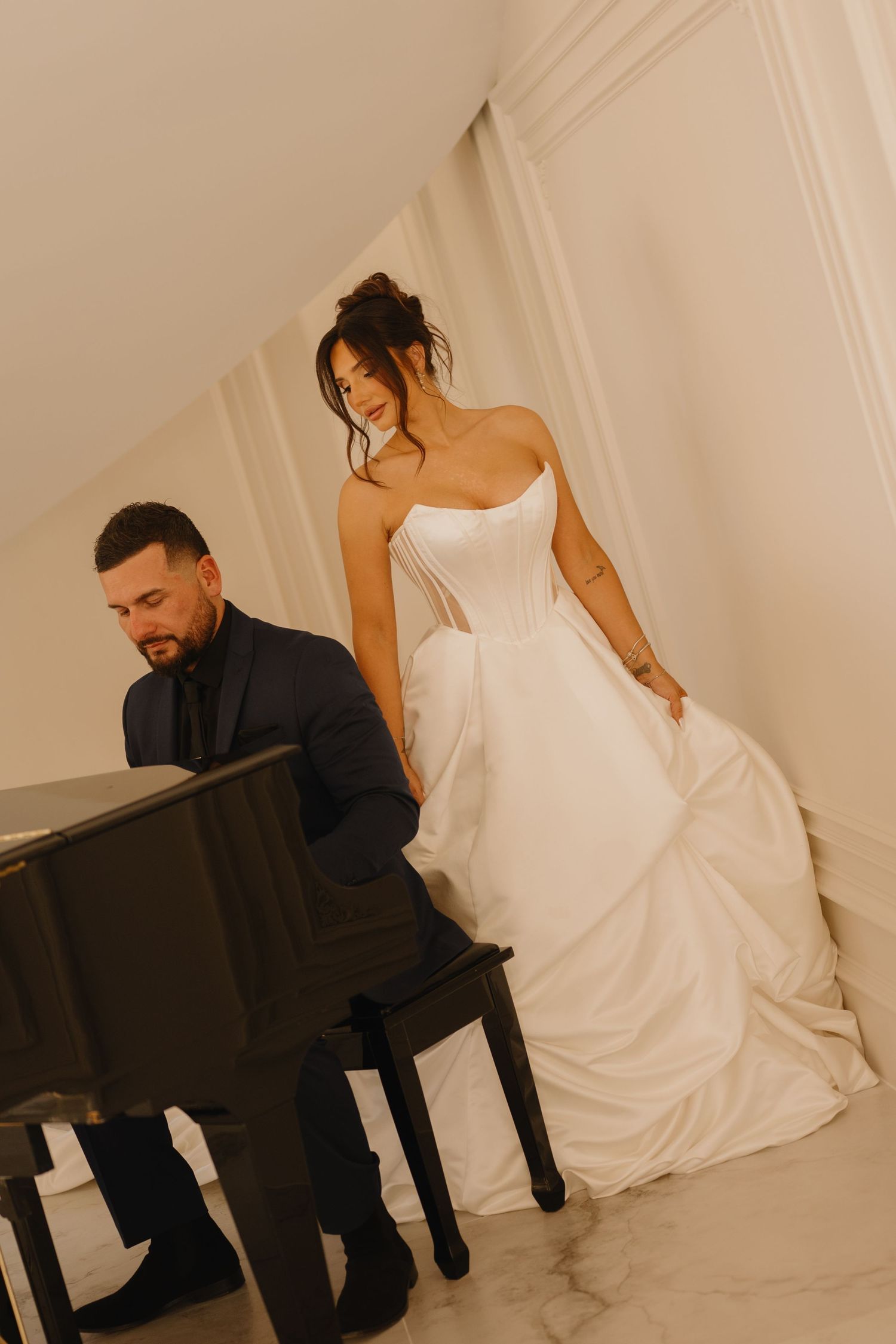 A groom in a dark suit plays a black piano while his bride in a strapless satin ball gown stands beside him in an elegant white room.
