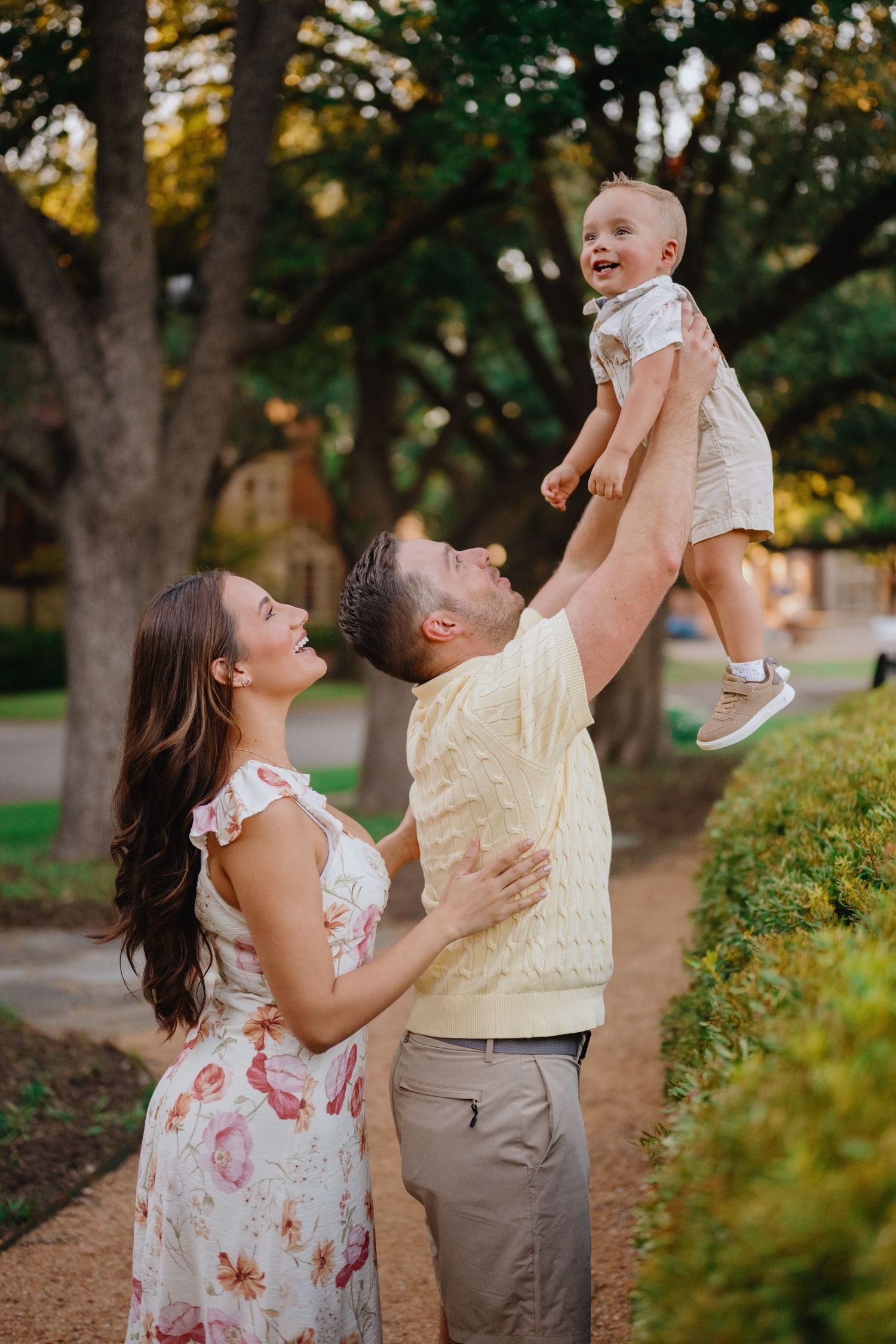 Smiling father lifts his giggling toddler into the air as the mother looks on in a tree-lined park.