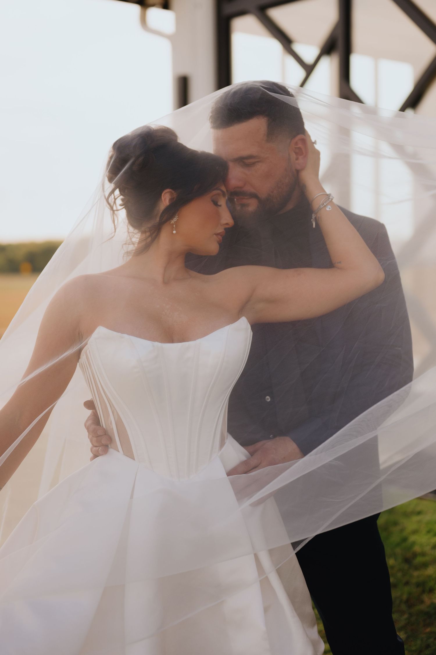 Couple embraces tenderly beneath a sheer wedding veil, the bride in a strapless corset gown, with a rustic structure behind.
