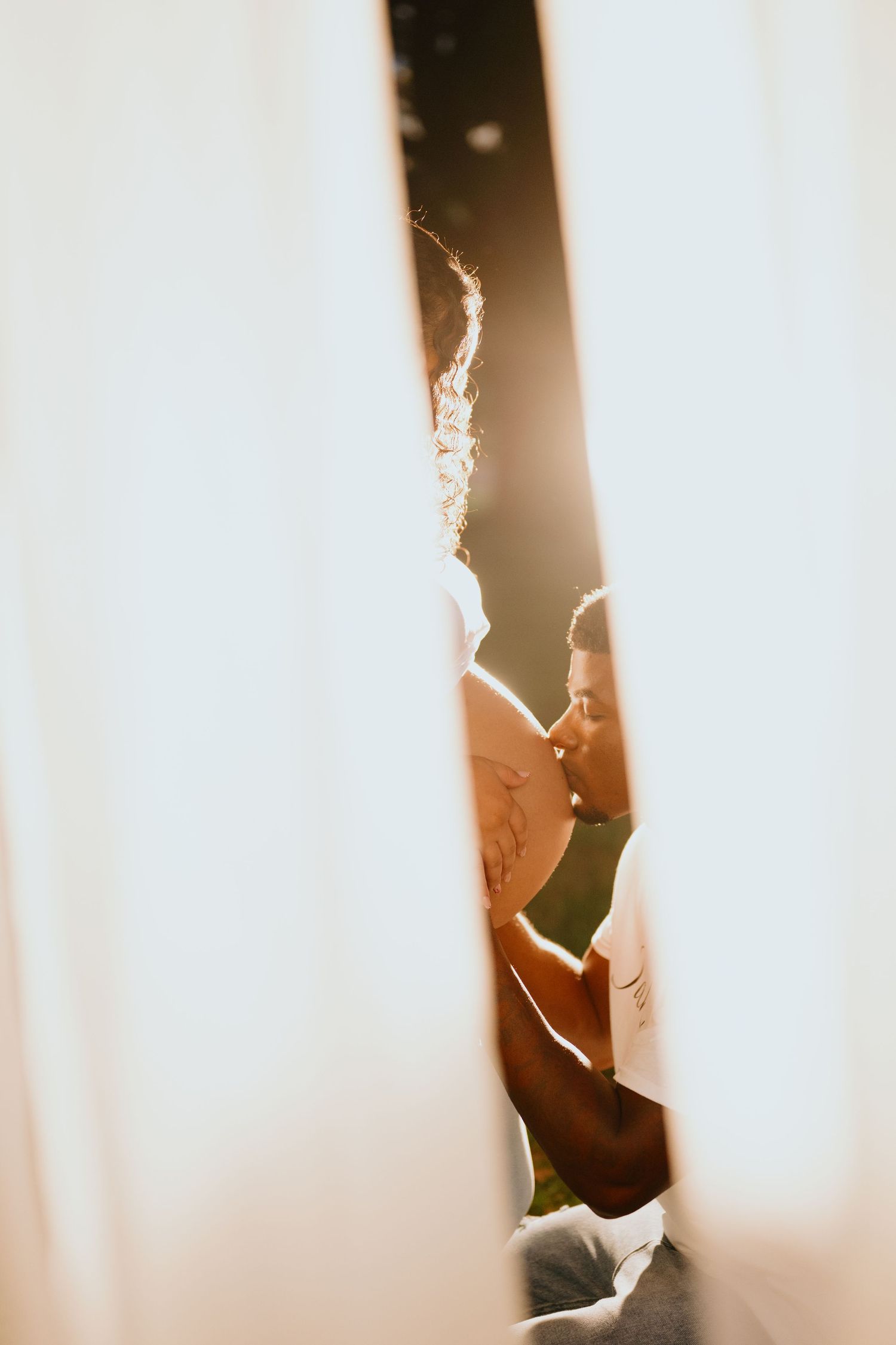 Soft sunlit maternity portrait of a man kissing his pregnant partner's belly, framed between flowing white curtains.