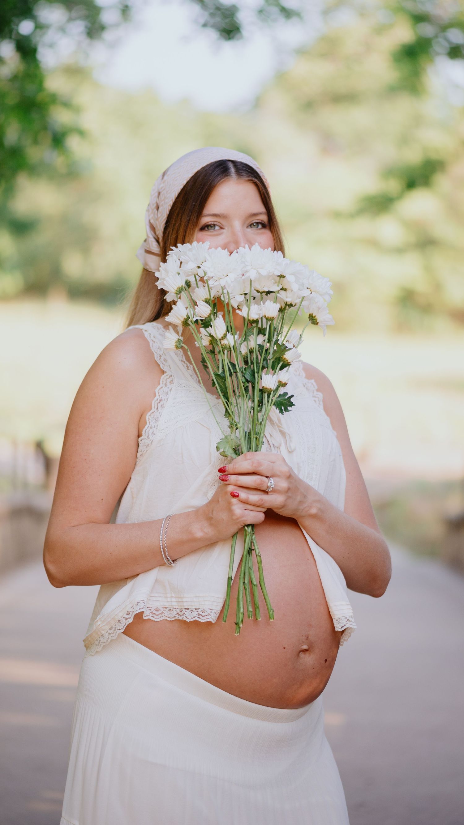 Pregnant woman in a white lace top and skirt holds a bouquet of white daisies in front of her face outdoors.