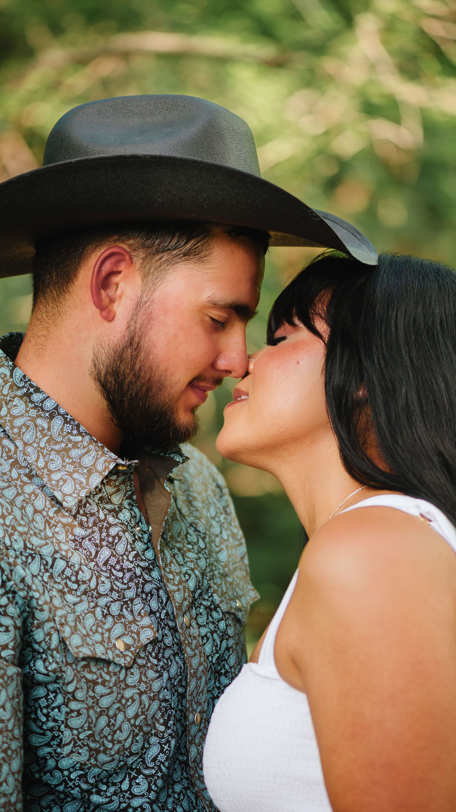 Couple about to kiss outdoors, the man in a cowboy hat and paisley shirt, the woman in a white top.