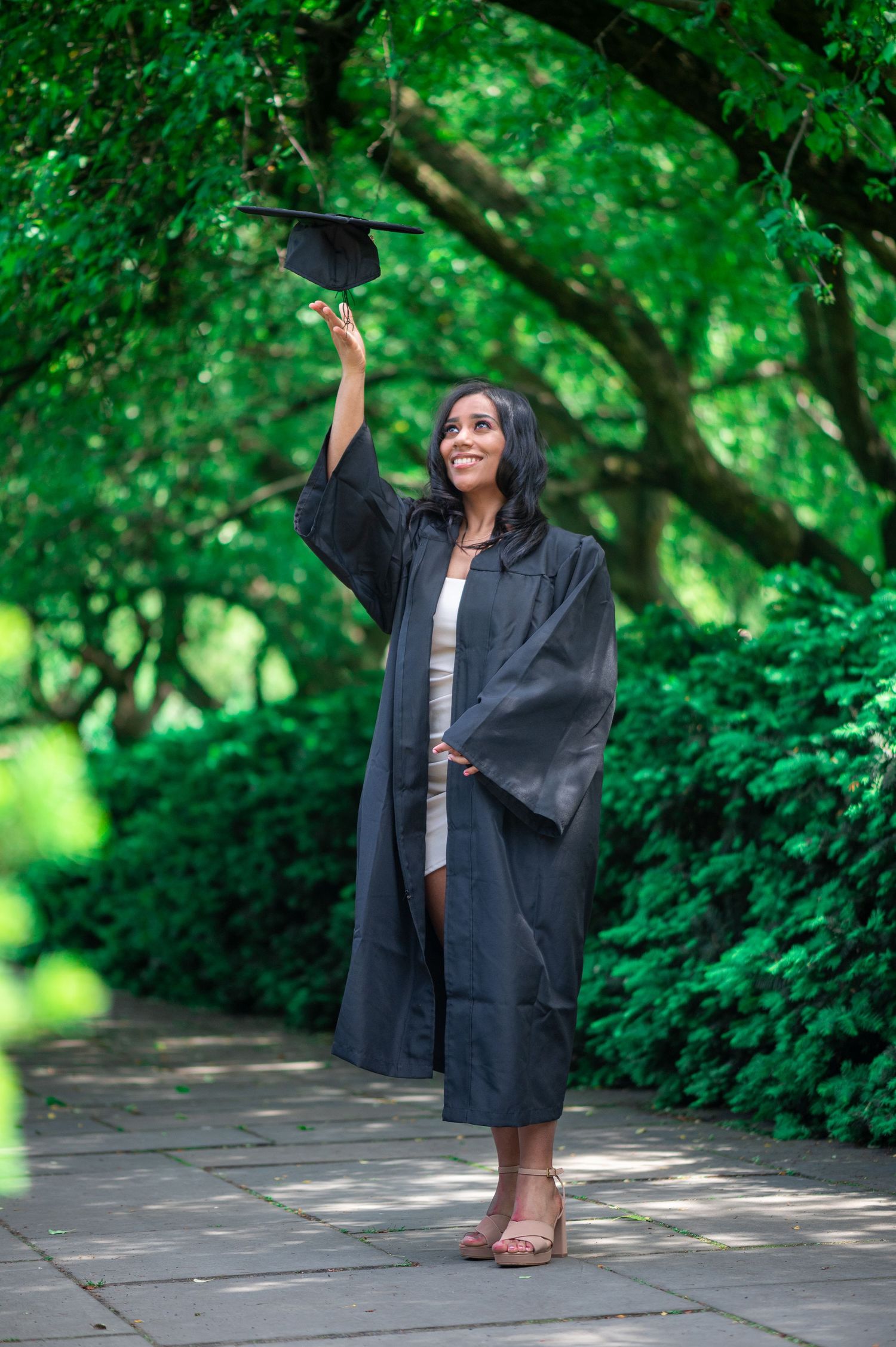 Smiling graduate in a black gown tosses her graduation cap into the air beneath arching green trees.