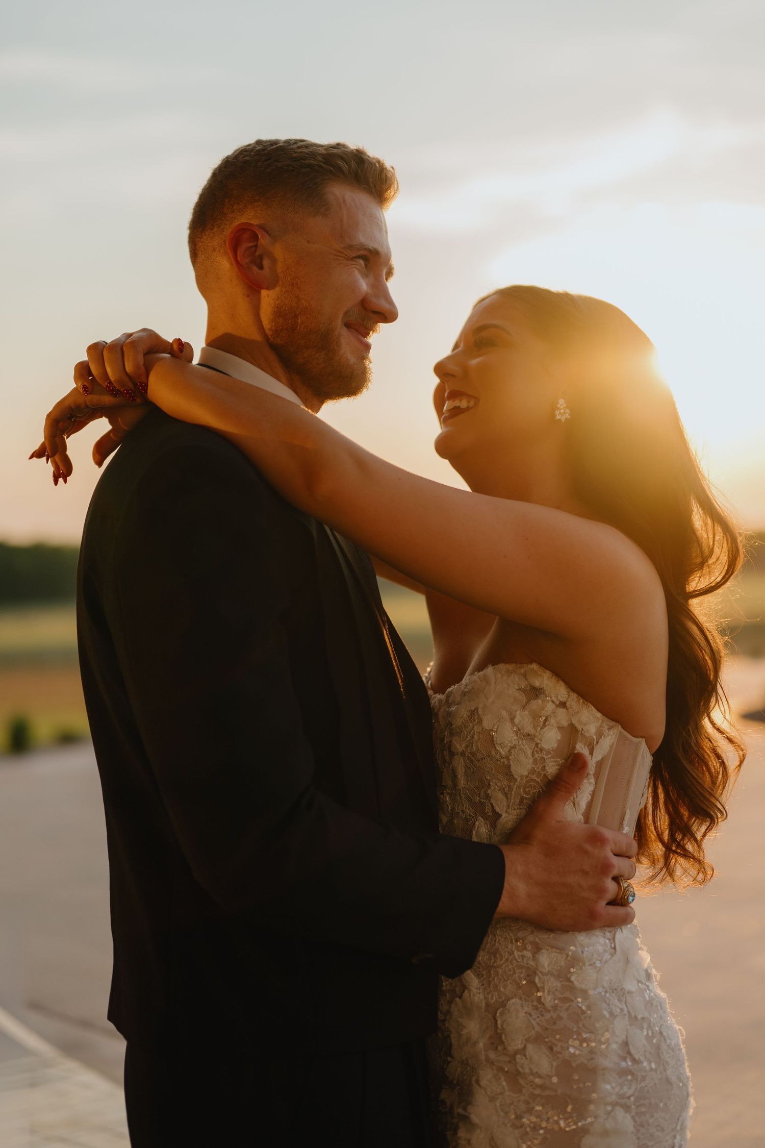 Bride in a beaded lace gown and groom in a dark suit laugh together in a warm embrace at golden hour sunset.