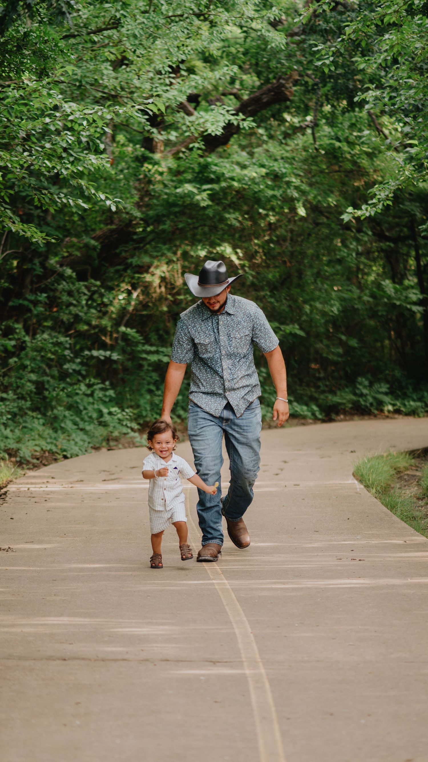 A father in a cowboy hat and jeans walks hand in hand with his toddler son along a paved path through lush green woods.