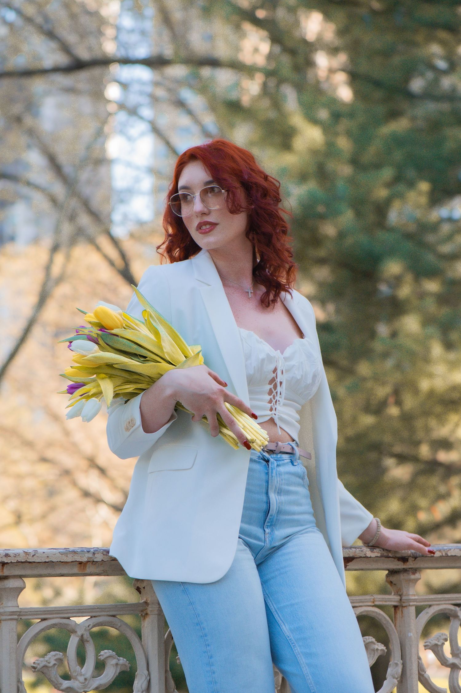 Red-haired woman in glasses, white blazer and light jeans holding yellow tulips while leaning on an ornate railing in a park.