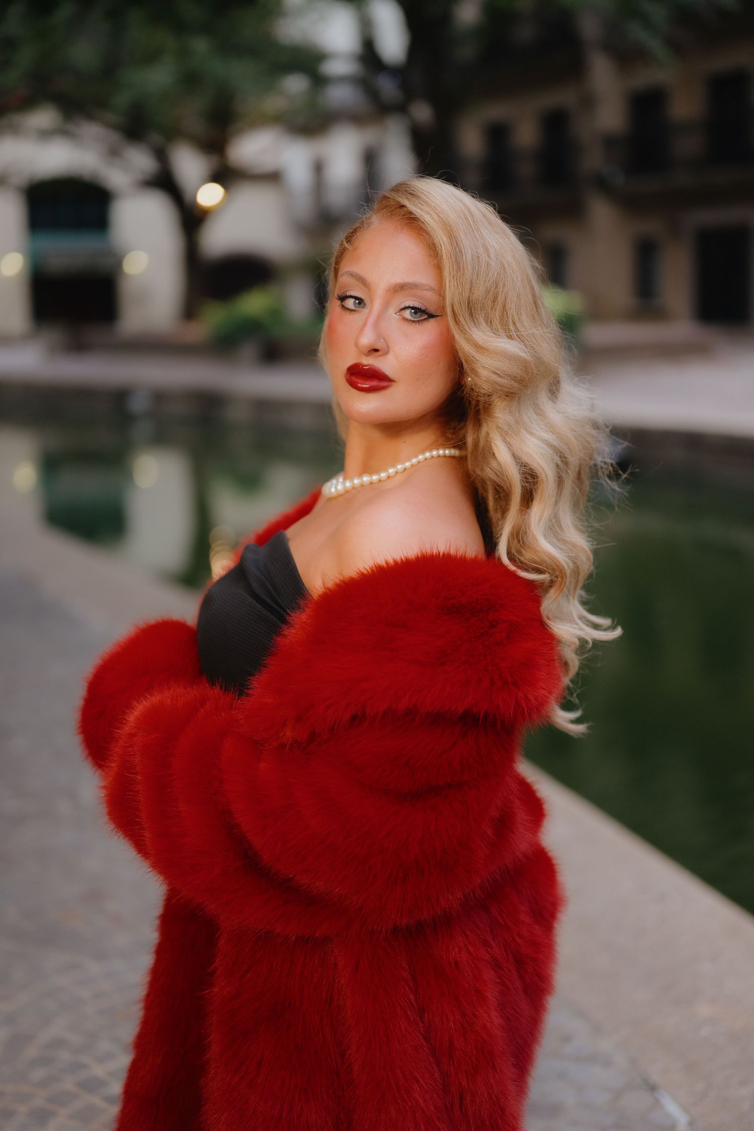 Blonde woman with wavy hair, red lipstick and a pearl necklace wearing a red faux fur coat beside a city canal.