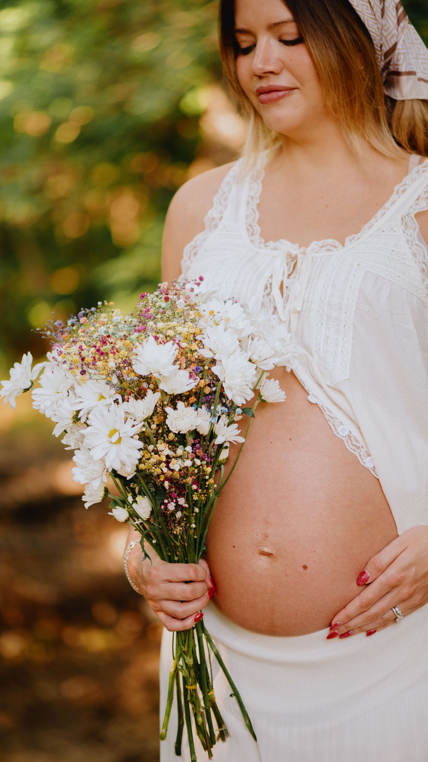 Pregnant woman in a white lace top and skirt holding a wildflower bouquet in a sunlit forest.