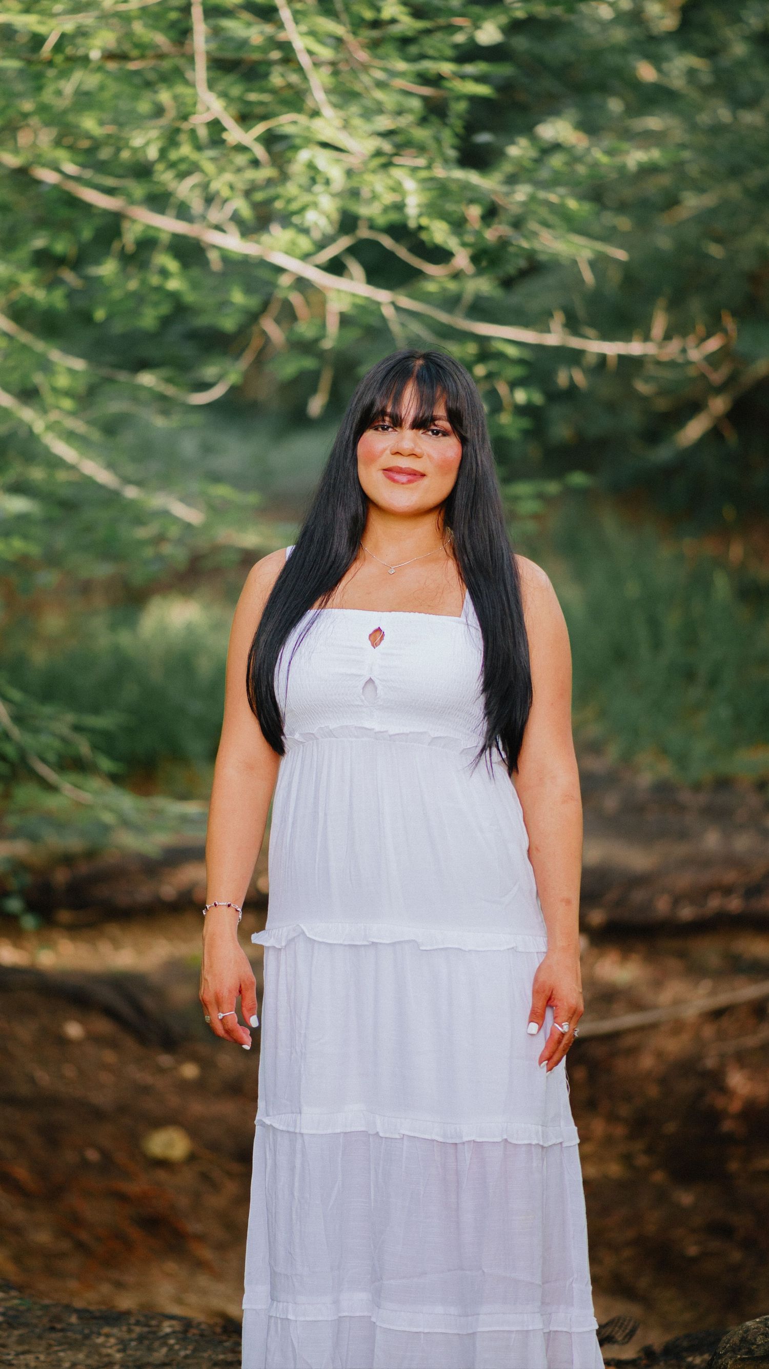 Woman with long dark hair and bangs wearing a white tiered sundress standing outdoors among green trees in a natural woodland setting.