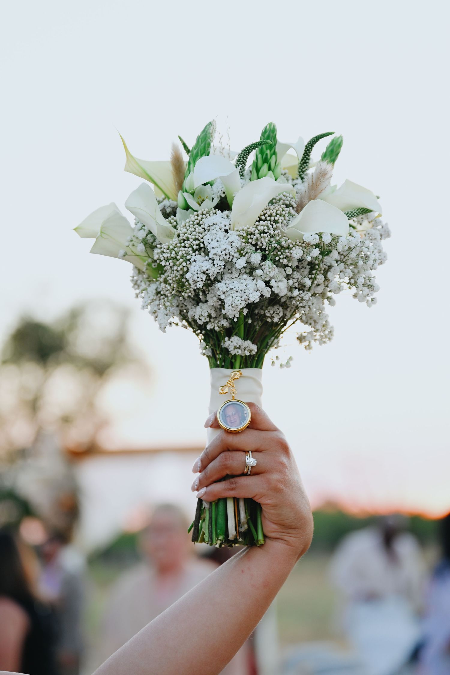 Hand holding a white wedding bouquet of calla lilies and baby's breath wrapped in ribbon with a photo charm as guests gather outdoors.