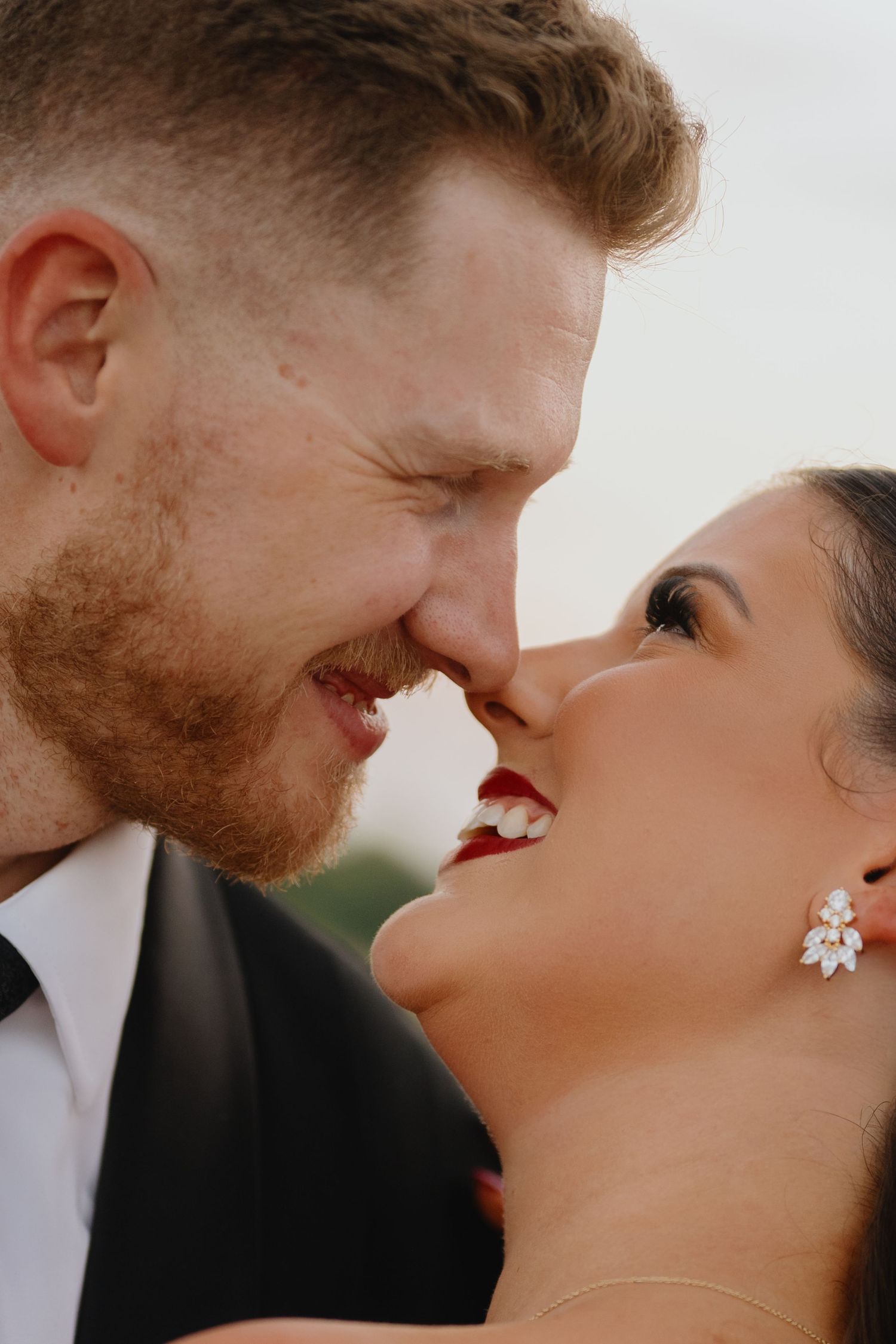 Close-up of a smiling bride and groom nose to nose, her red lipstick and crystal drop earring catching the soft light.