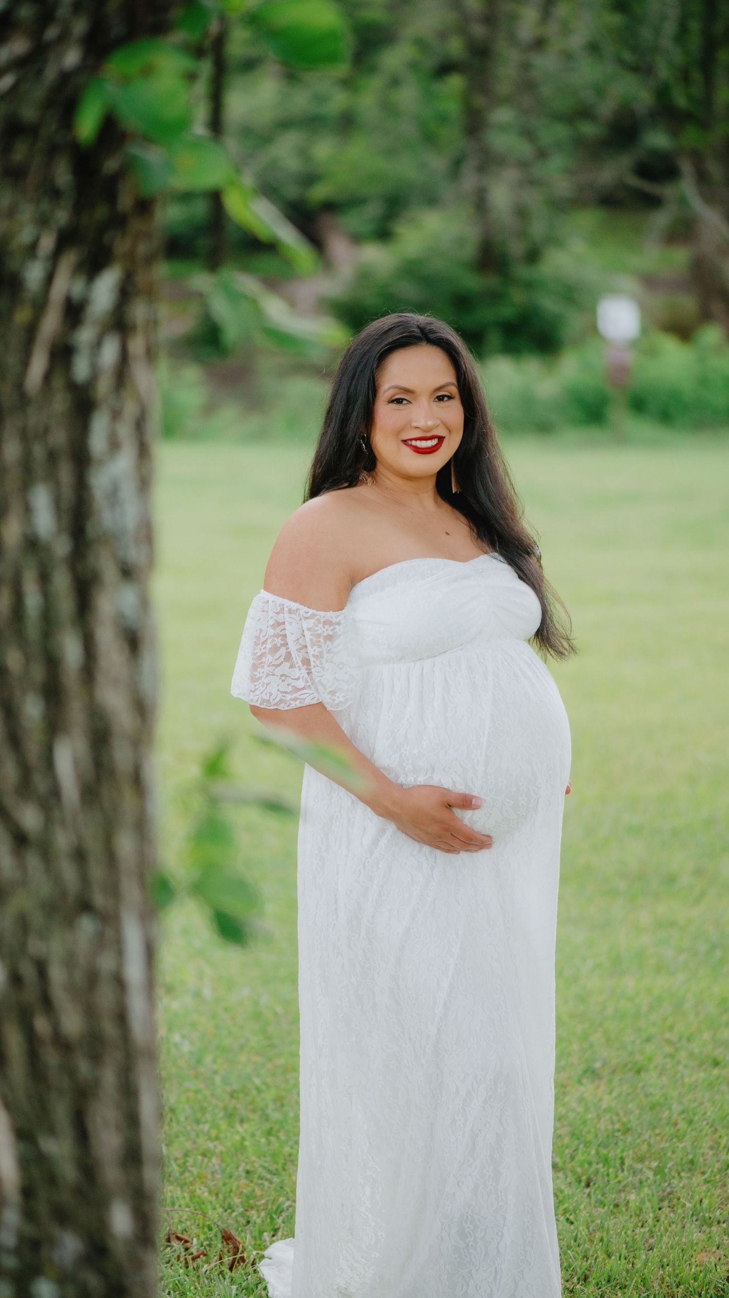 Pregnant woman in a white off-the-shoulder lace maternity gown stands on a green lawn beside a tree, hand on her belly.