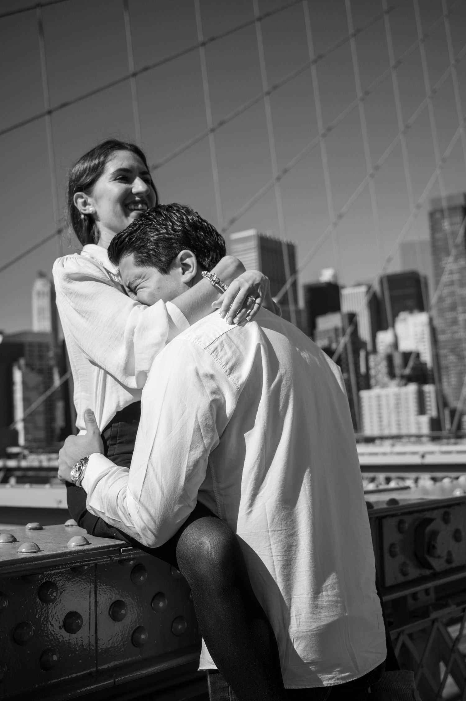Black and white photo of a couple embracing on the Brooklyn Bridge, the woman laughing with a ring on her hand and city skyline behind.