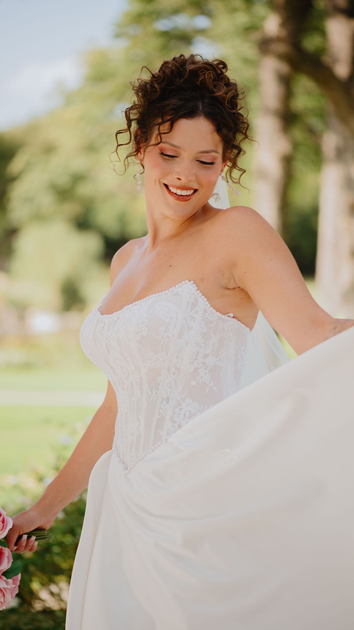Smiling bride with curly updo and lace bodice wedding dress twirls her skirt while holding pink flowers outdoors.