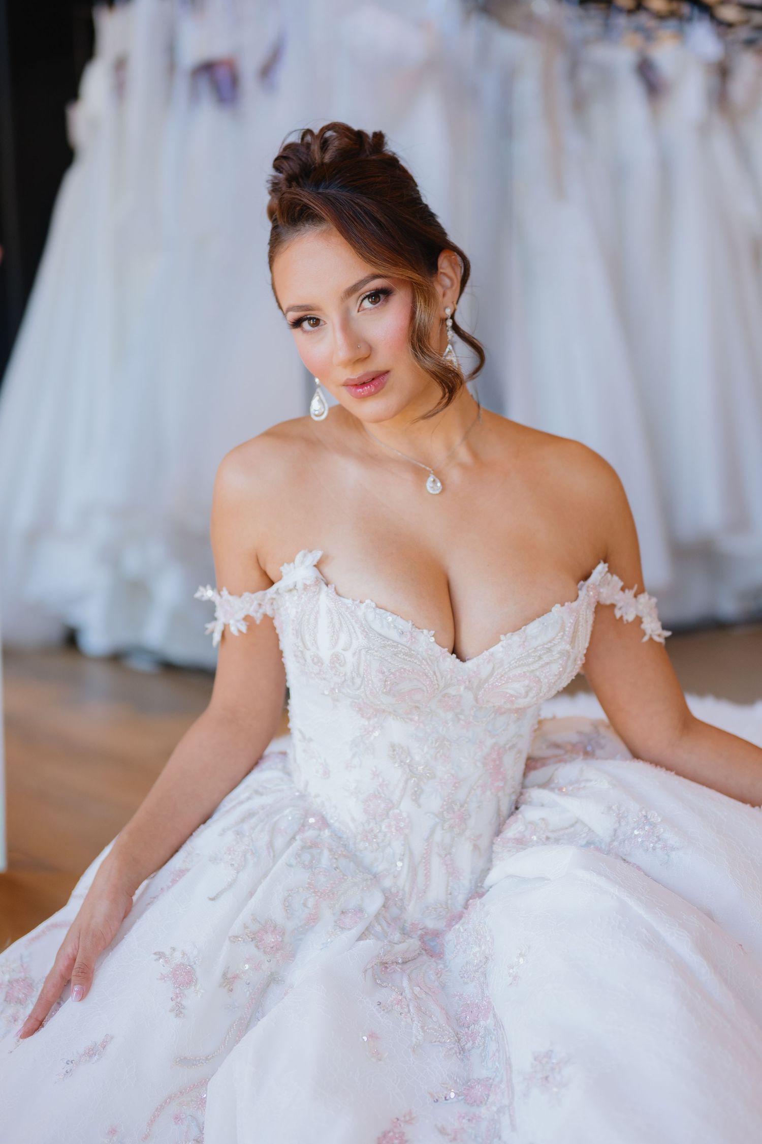 Close-up of a smiling bride in a lace off-the-shoulder wedding gown with drop earrings and an updo, seated before racks of bridal dresses.
