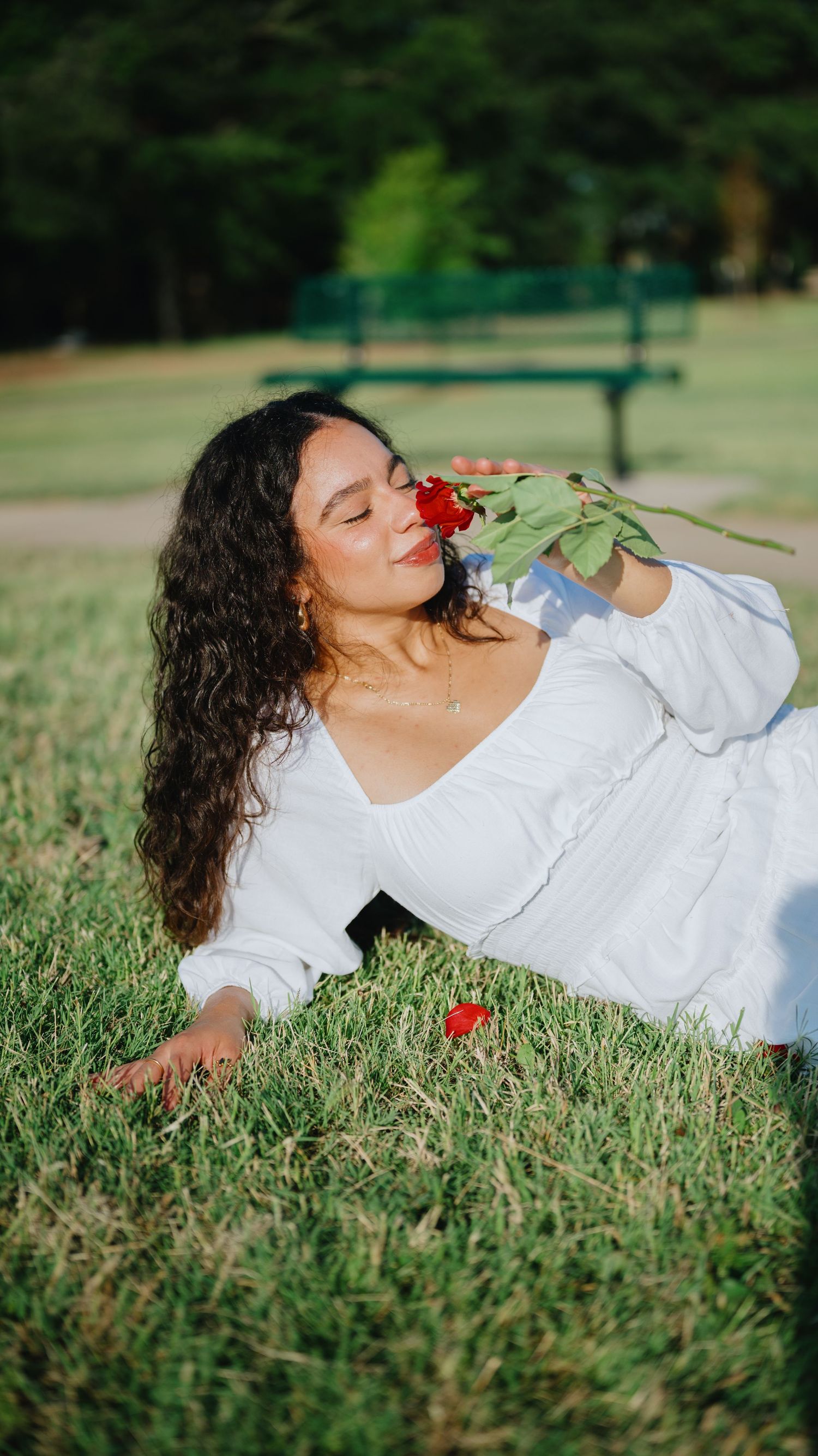 Woman with long curly hair in a white dress relaxing on green grass while smelling a red rose in a sunny park.