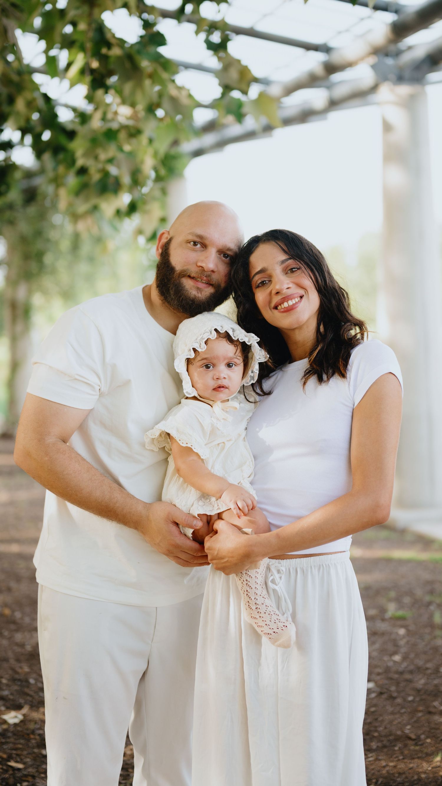 A smiling couple in white outfits stand together under a leafy pergola holding their baby girl in a cream lace dress and bonnet.