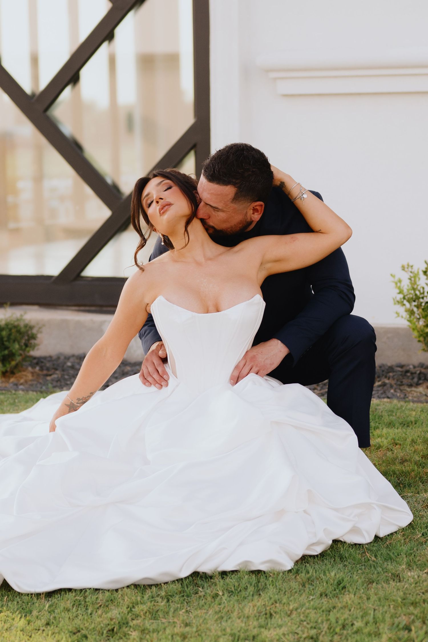 Groom in a navy suit kisses a bride's neck as she sits in a full white ballgown on a sunny green lawn.