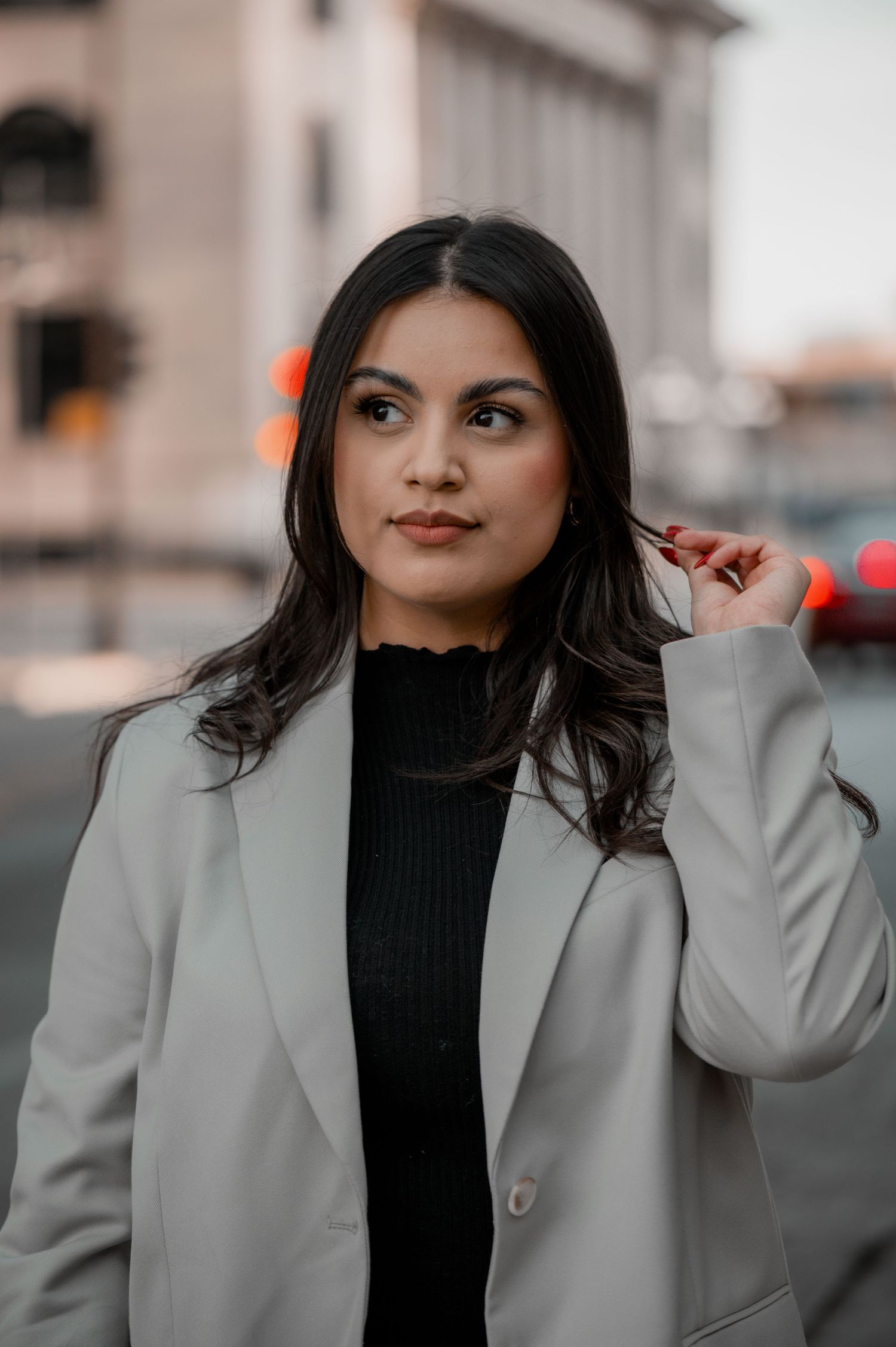 Close-up portrait of a woman with long dark hair in a gray blazer and black top, looking aside on a city street.