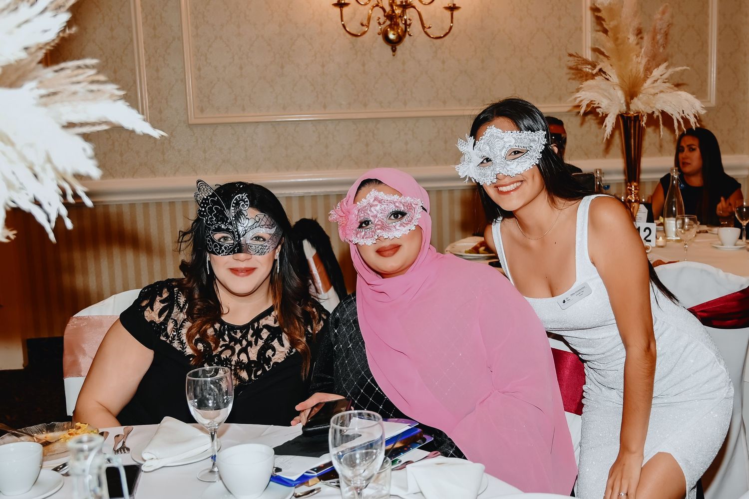 Three smiling women in decorative masquerade masks pose together at a table at an elegant formal party.