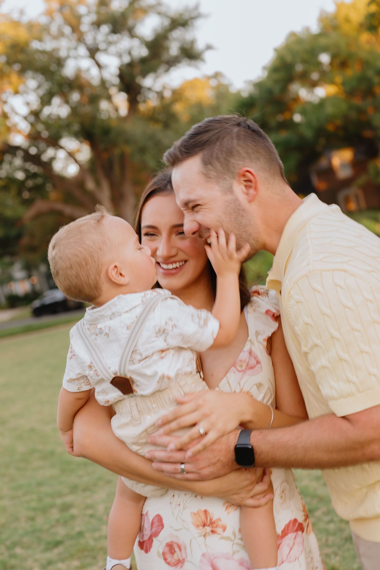 Smiling mother and father hold and nuzzle their baby in a sunny park during a warm family photo session.