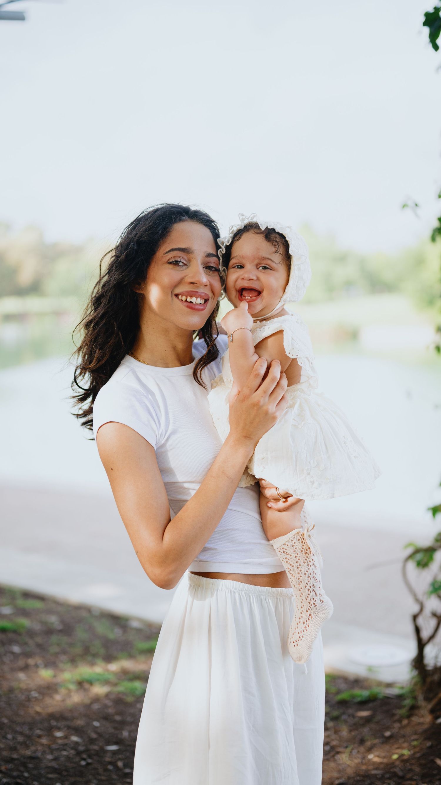 Joyful mother in white outfit holds her laughing baby girl in a lace bonnet beside a calm lake.