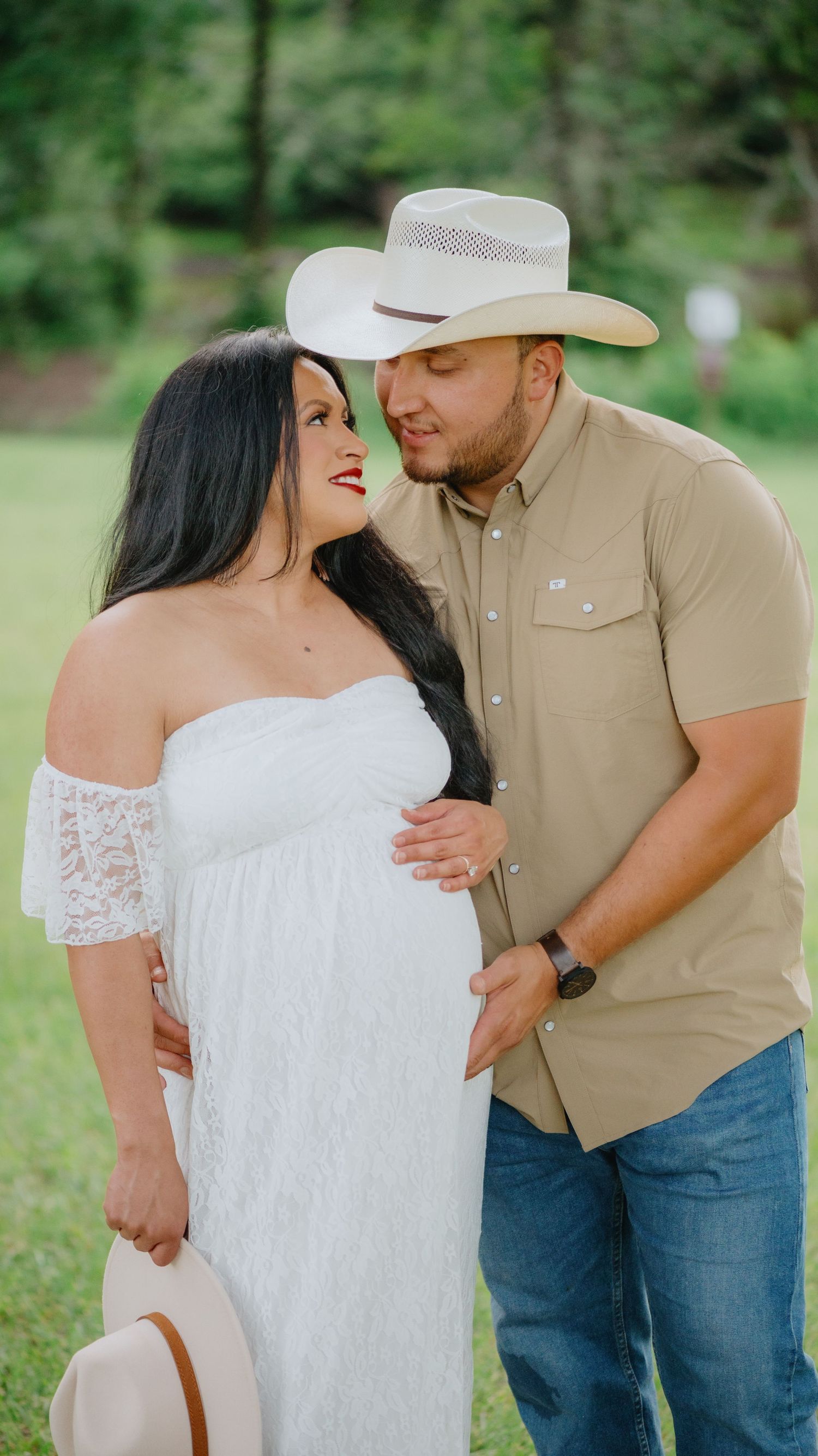 Expecting couple embraces in a grassy park, she in a white lace dress and he in a cowboy hat and tan shirt, hands on her belly.