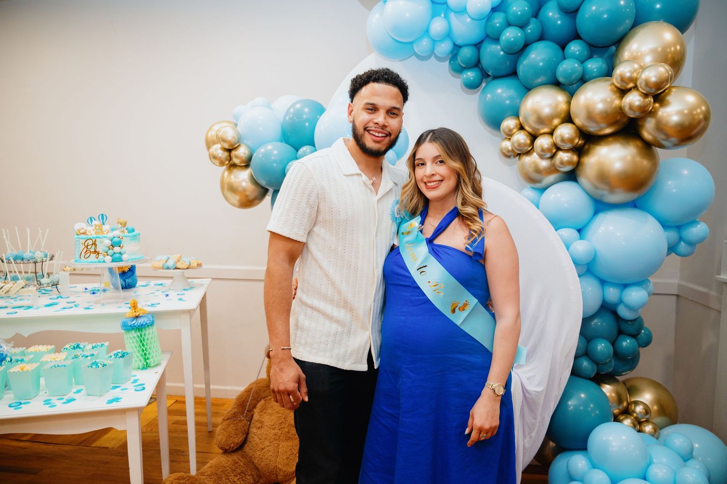 Smiling couple poses at a baby shower with blue and gold balloons, a dessert table, and a teddy bear.