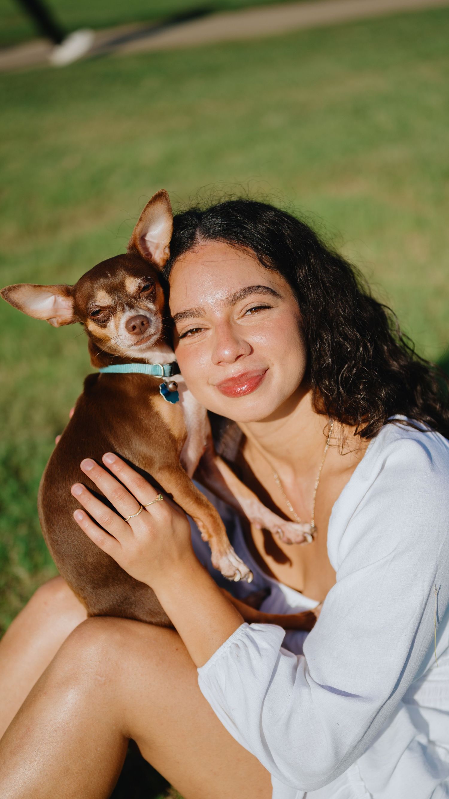 A smiling woman with curly hair in a white top hugs her small brown chihuahua on a sunlit grassy lawn.