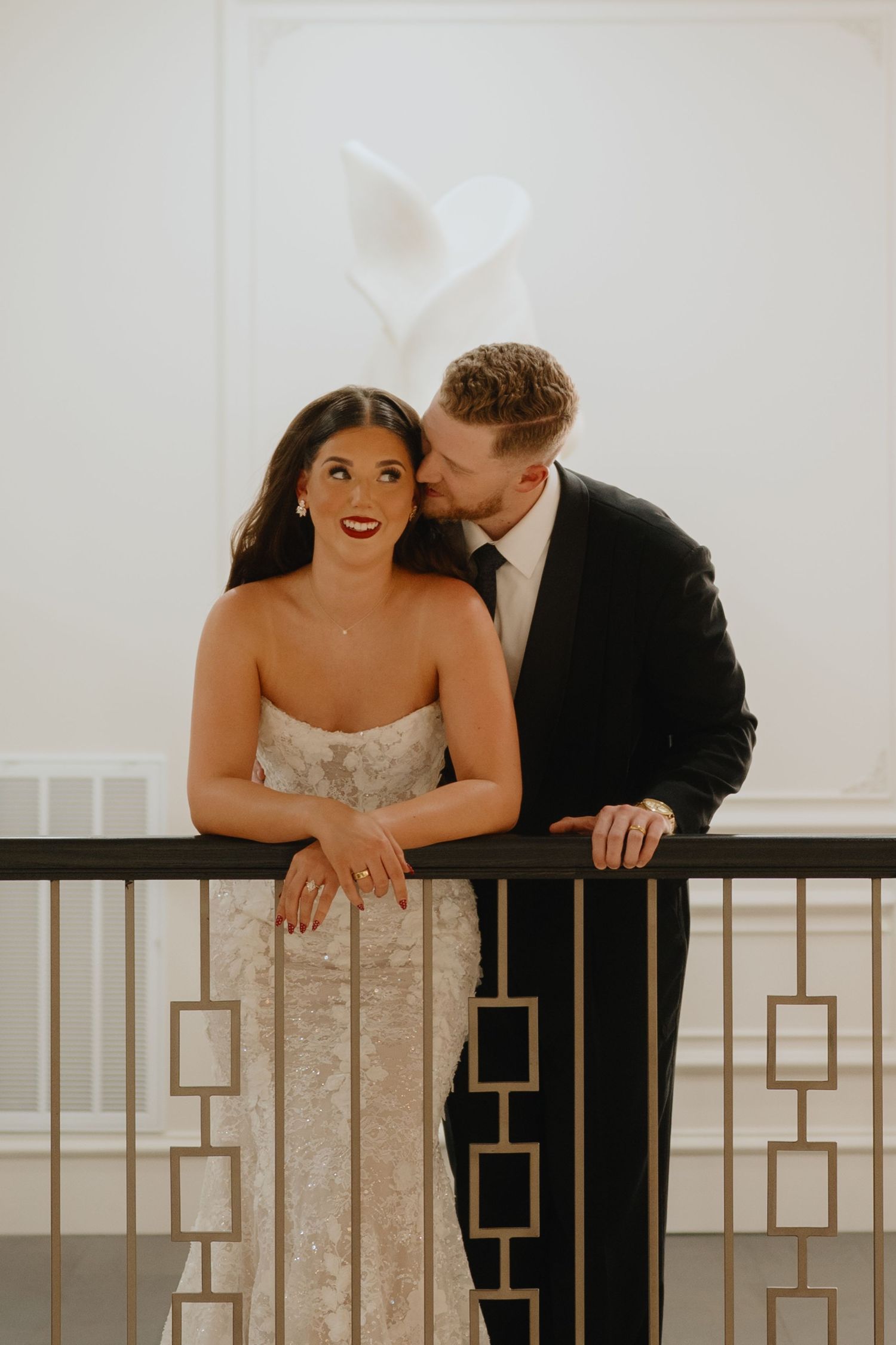 Groom in a black suit kisses his smiling bride's cheek as they lean on a gold-accented railing at their wedding venue.