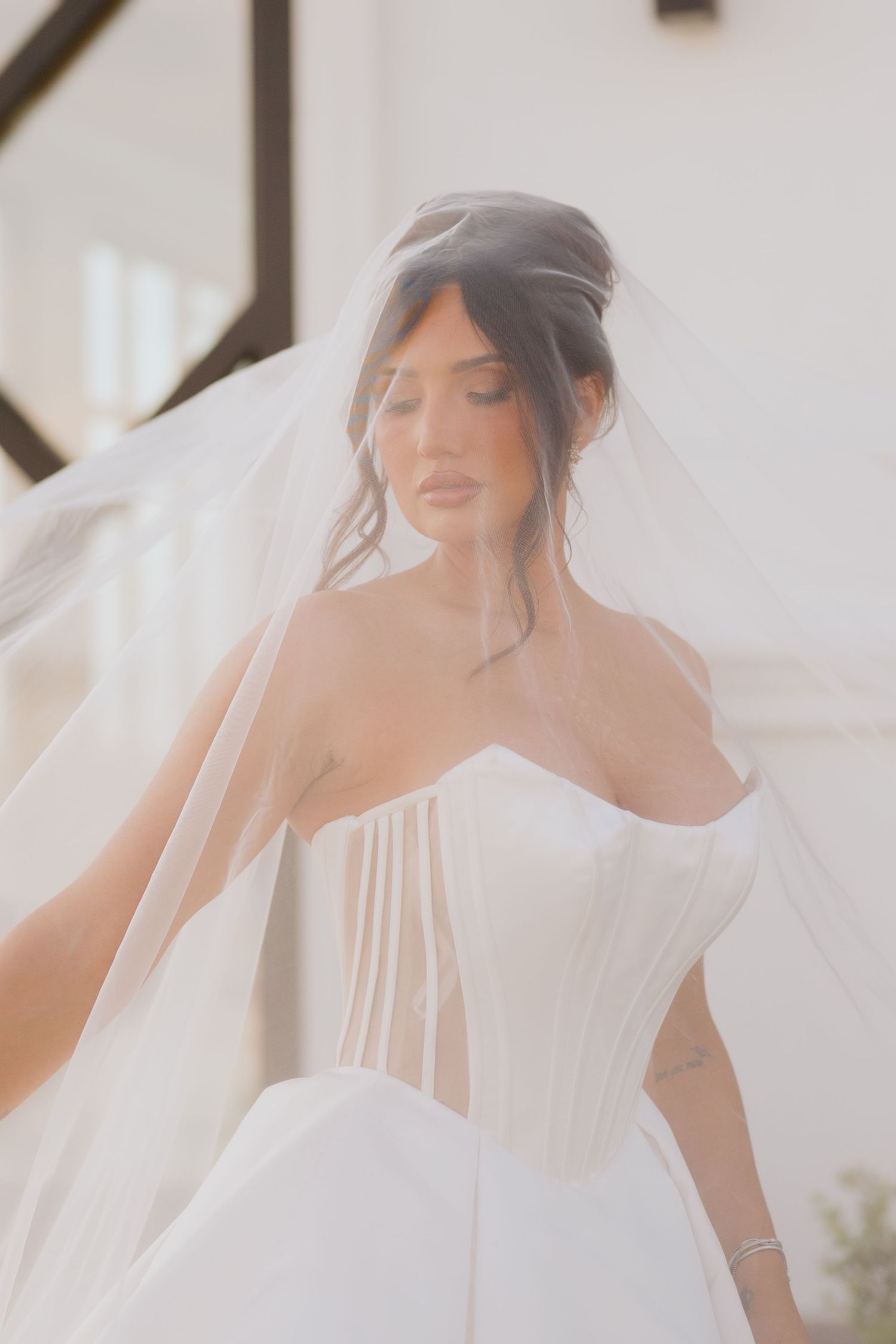 Bride in a sheer-paneled strapless corset gown gazes down beneath a flowing tulle veil in soft natural light.