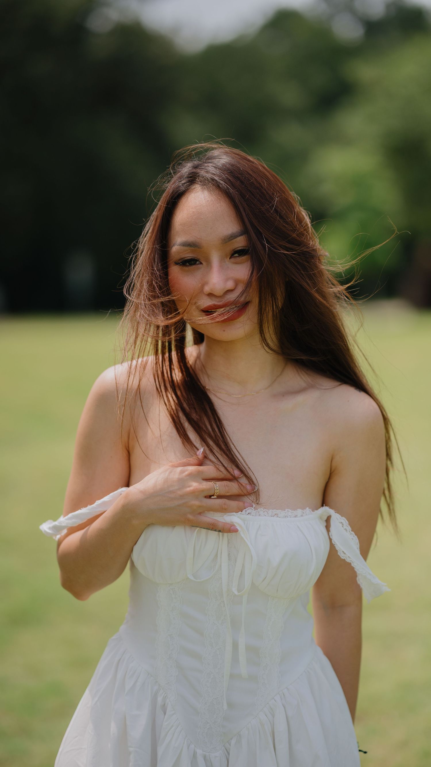 A woman with long windblown hair in an off-the-shoulder white dress smiles softly in a green park.