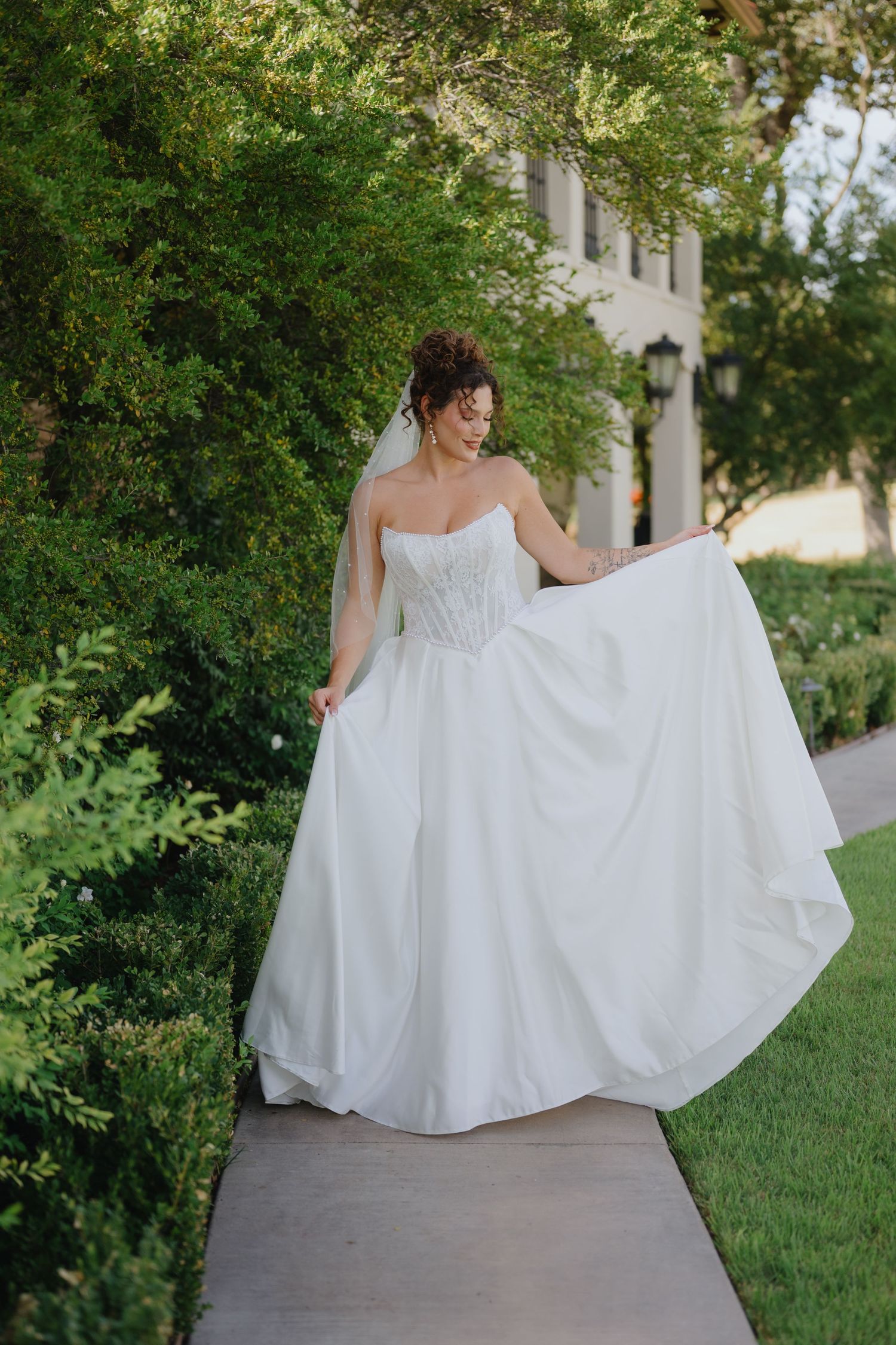 Bride in a flowing white satin gown and veil twirls her skirt along a sidewalk lined with lush green hedges.