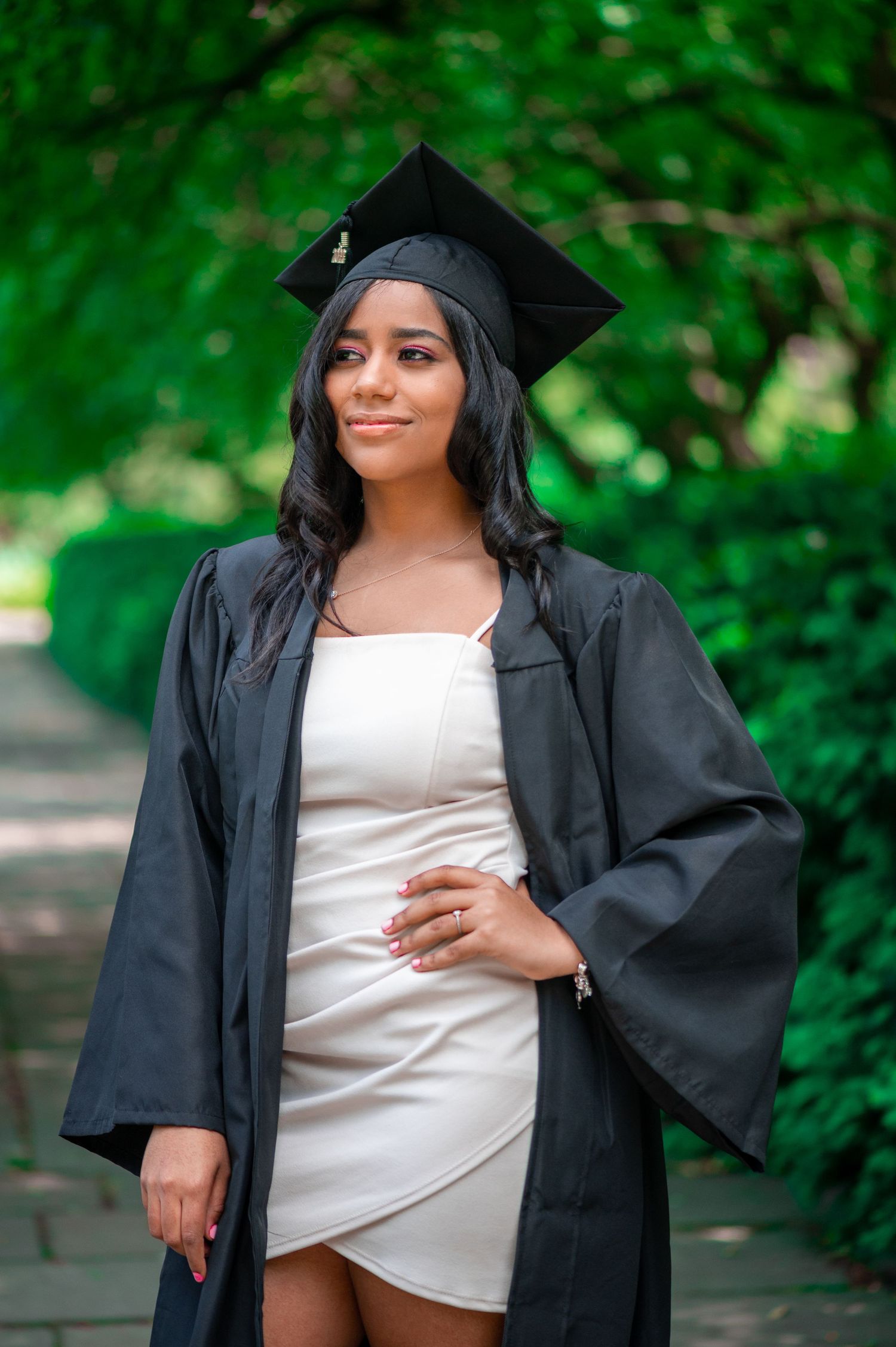 Graduate in a black cap and gown over a white dress stands on a tree-lined path in a lush green park.