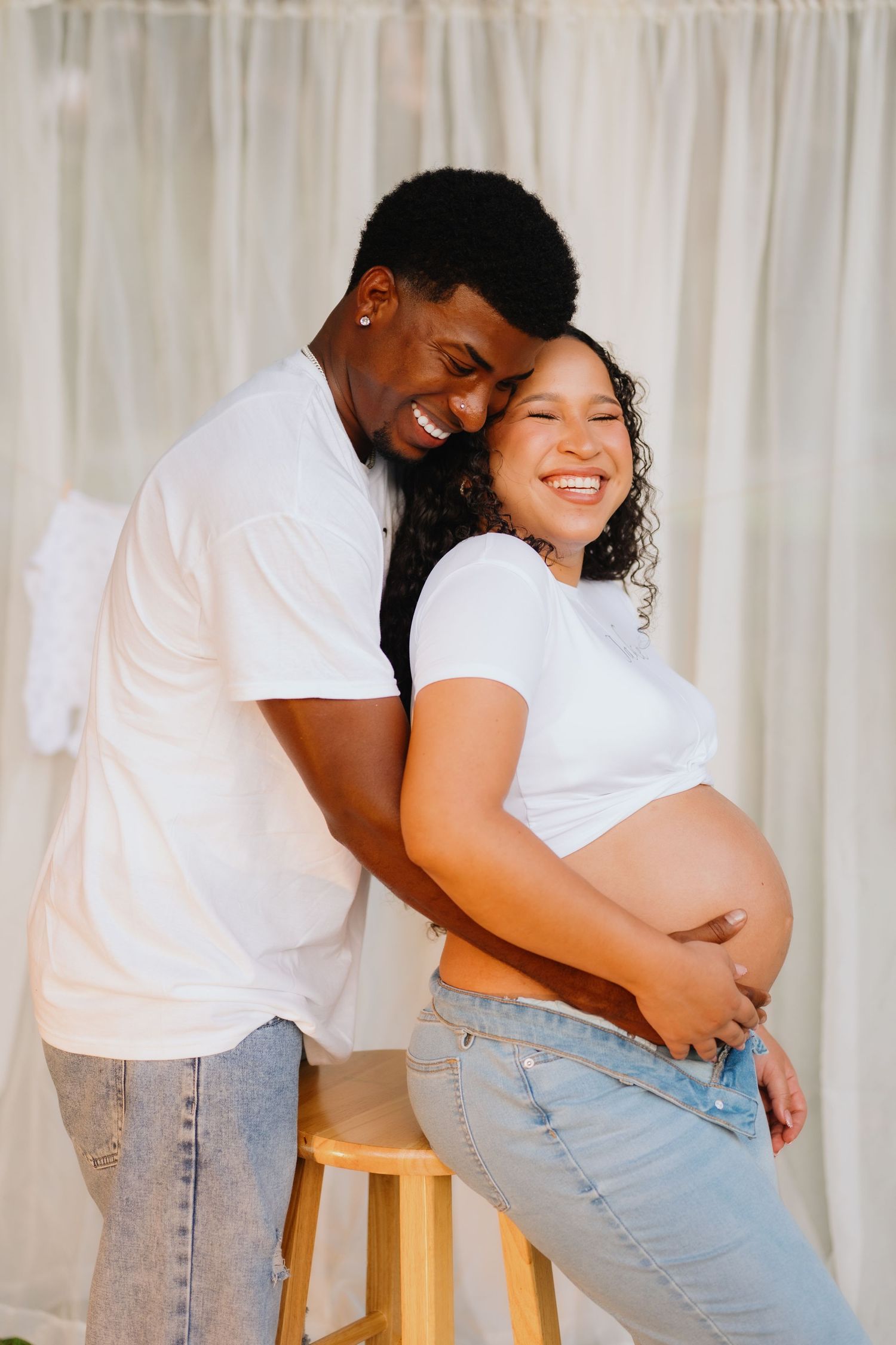Smiling couple in white tees and jeans embrace and cradle her baby bump during a maternity photo session.