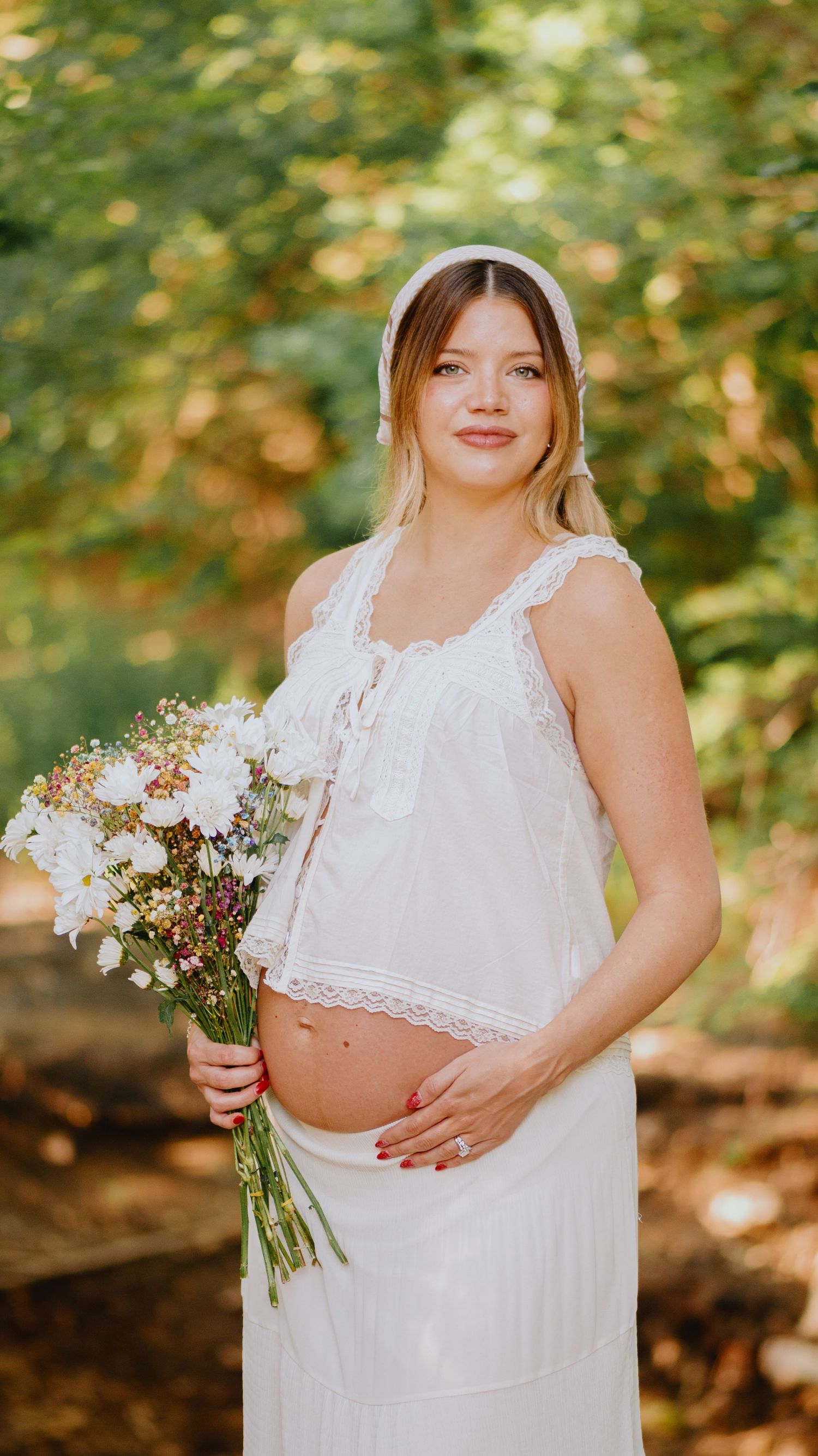 Pregnant woman in a white lace-trimmed top and skirt holds wildflowers while posing in a sunlit wooded setting.