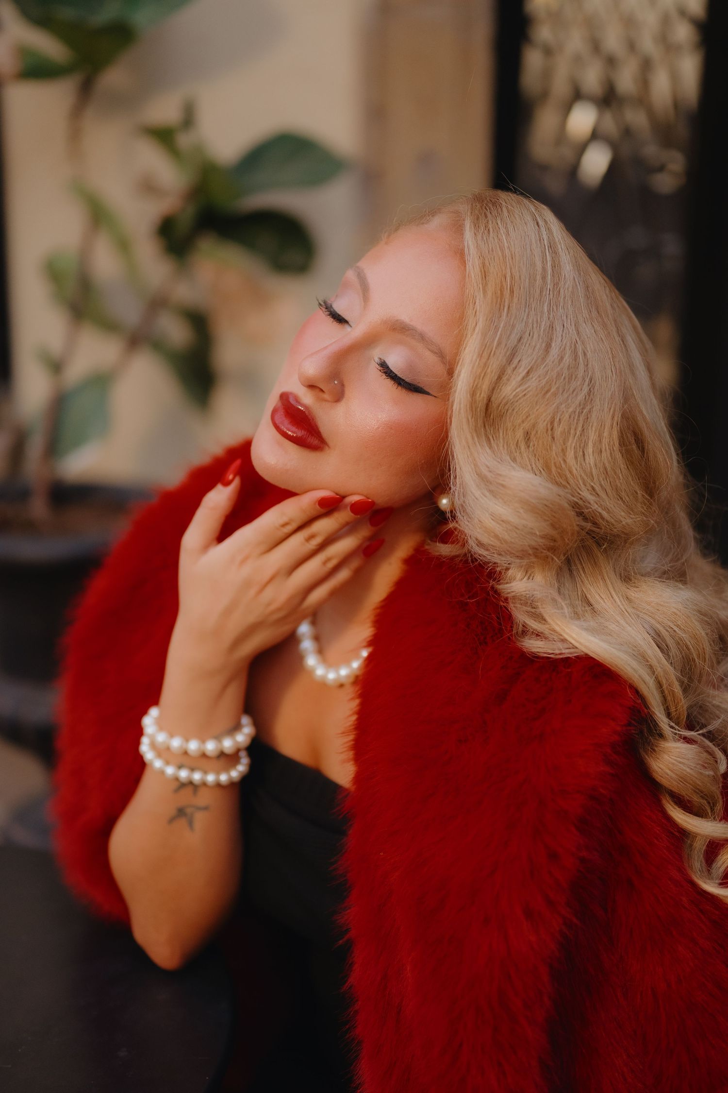 Glamorous blonde woman in a red fur wrap and pearl bracelets closes her eyes and touches her neck beside a green plant.
