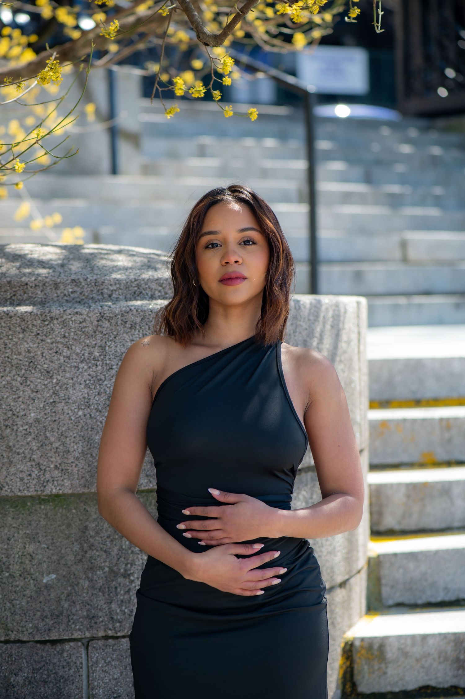 Woman in a black one-shoulder dress posing with hands on her stomach beside stone steps and a tree with yellow spring blossoms.