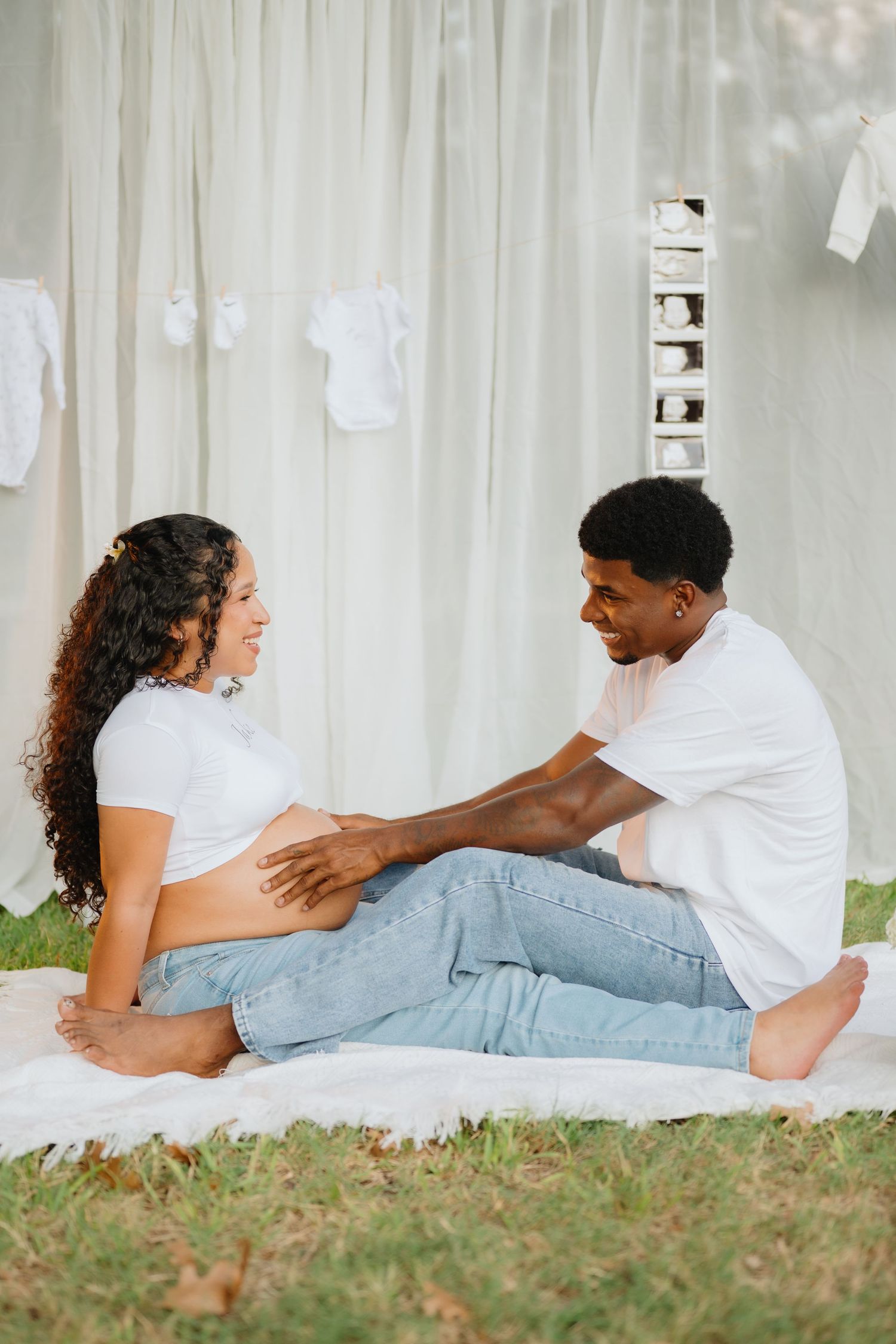 Smiling couple sits on a white blanket on the grass as the man touches his partner's pregnant belly, baby clothes hanging behind.