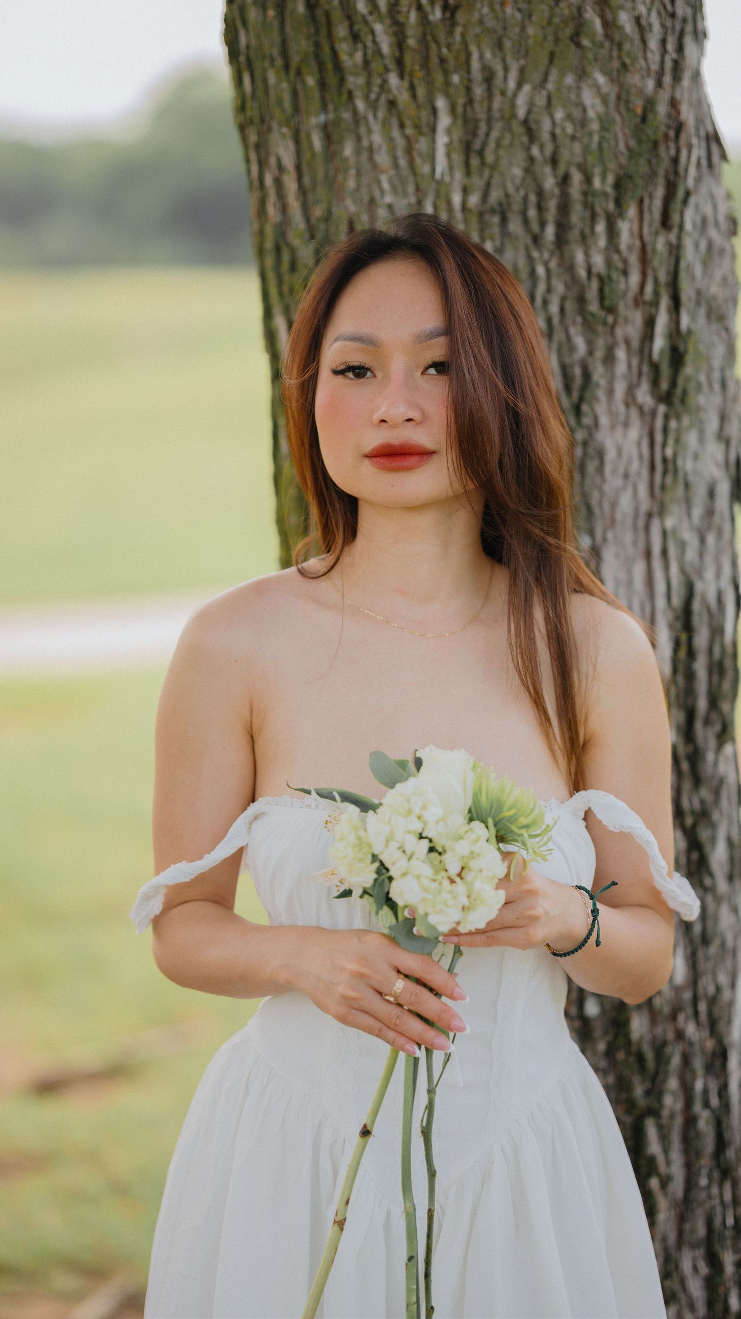 Woman in a white strapless dress holding a white floral bouquet while standing beside a tree in a grassy park.
