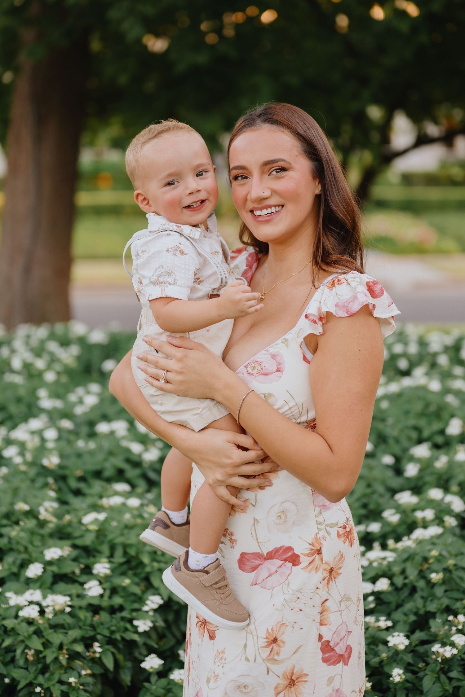 Smiling mother in a floral dress holds her toddler son in front of white blooming flowers at a sunny park.