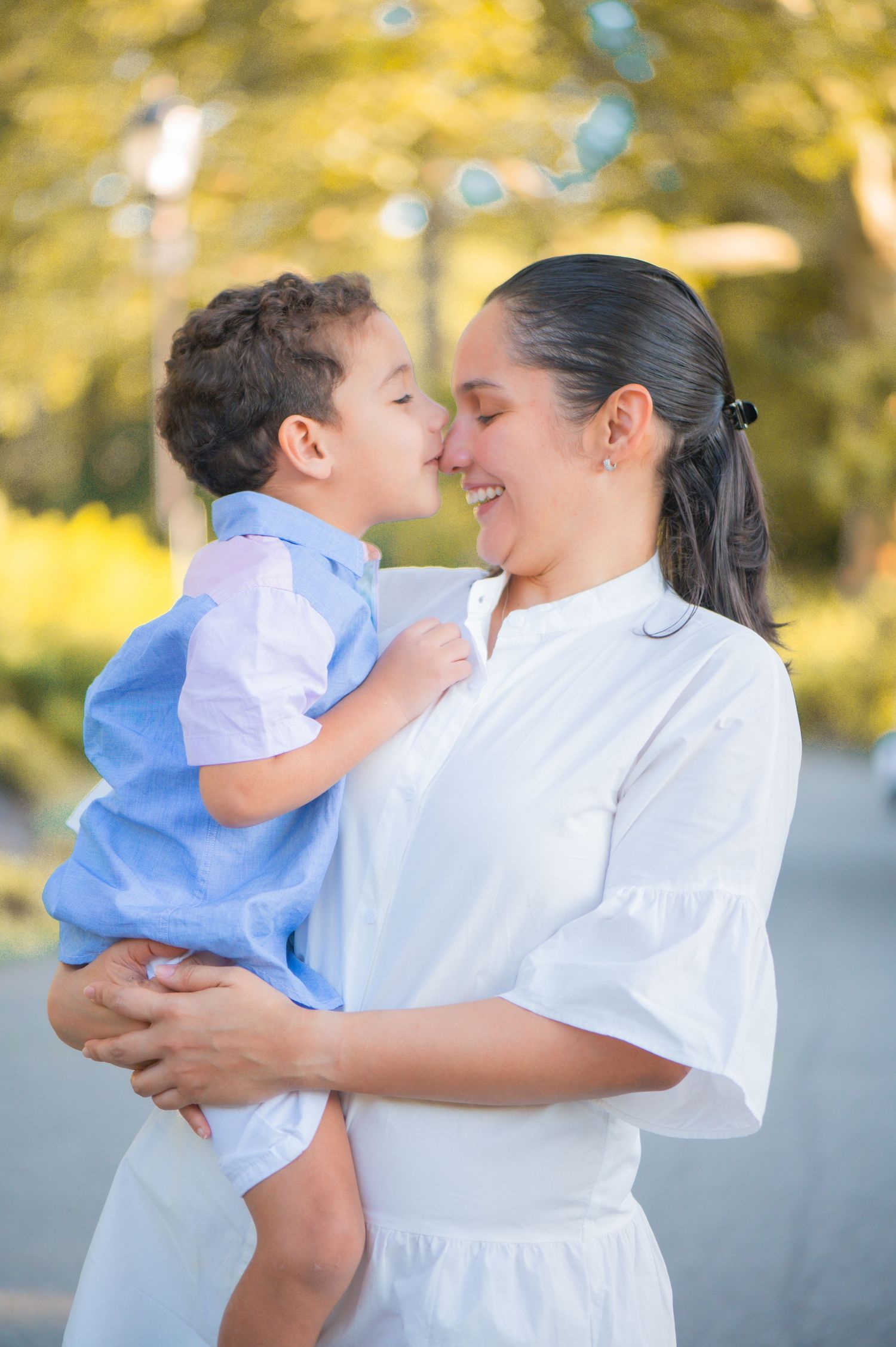A smiling mother in a white dress holds her young son in a blue shirt as he touches noses with her in a sunlit park.