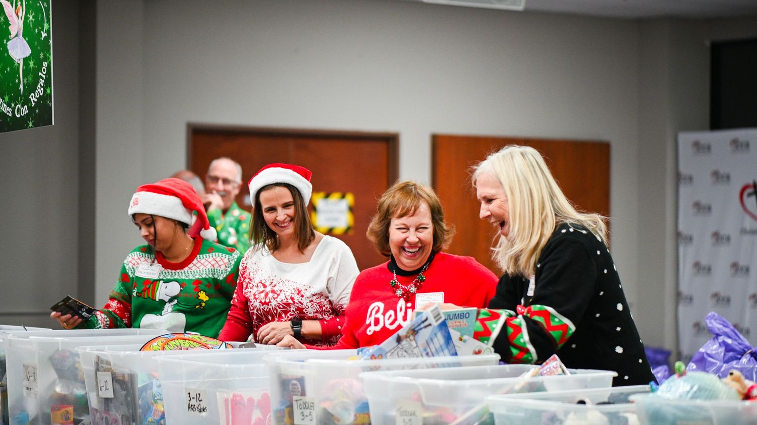Four volunteers in Christmas sweaters and Santa hats sort supplies in plastic bins at a holiday charity event.
