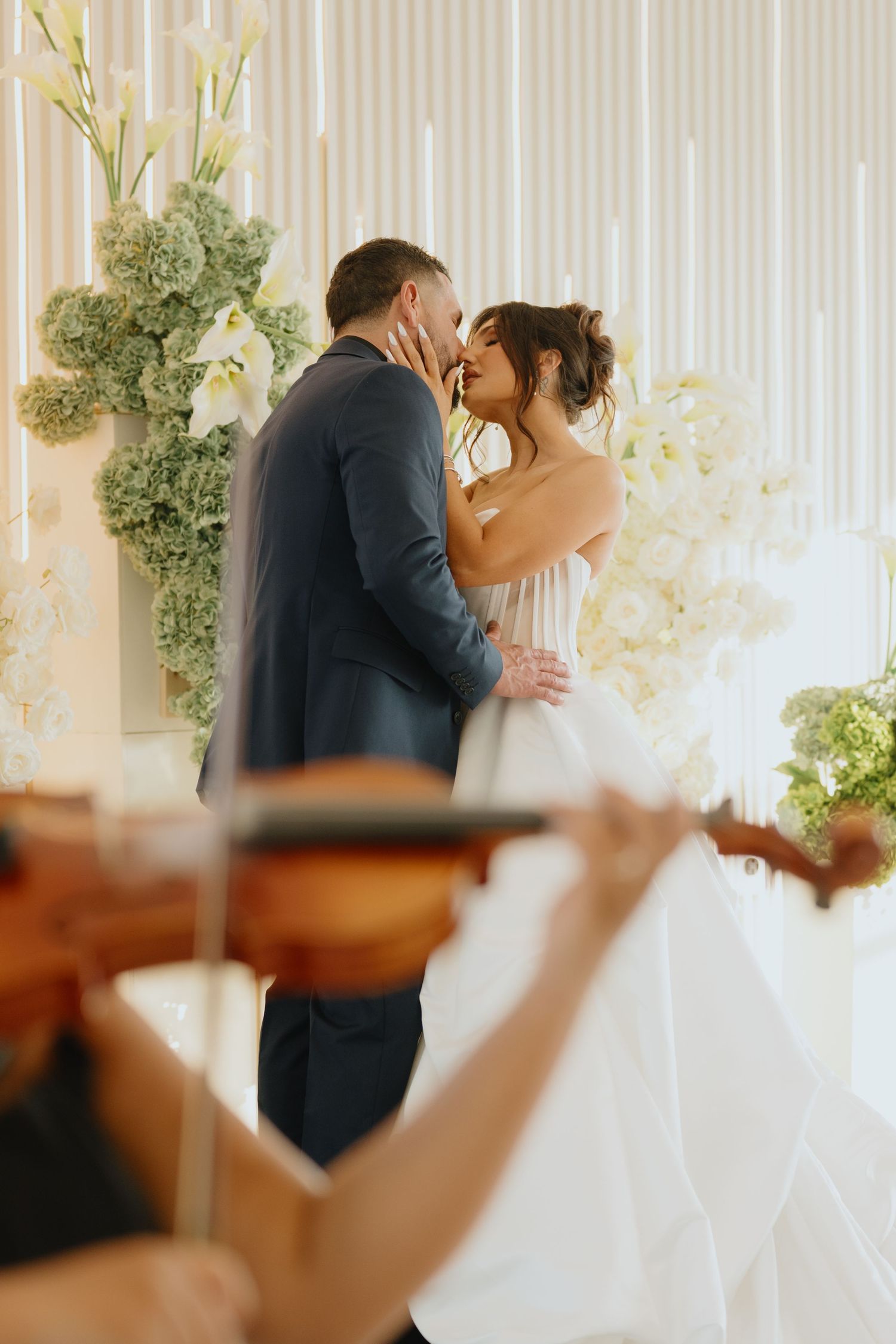 Bride and groom share a kiss among white floral arrangements while a violinist plays in the blurred foreground.